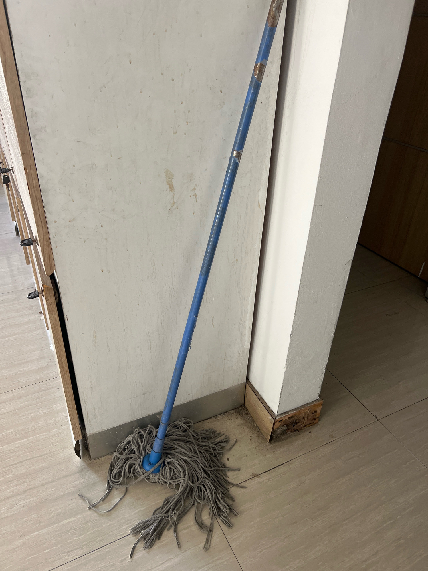 A mop with a blue handle and gray string head rests on a light-colored tiled floor, leaning against a white wall next to a wooden cabinet. The wall has some scuff marks, and the baseboard where it meets the floor is damaged. The floor tiles are arranged in a grid pattern. The scene appears to be indoors, possibly in a hallway or utility area. No people are present in the image. The overall impression is one of an ordinary, utilitarian space. The image does not provide any information about the city of Yogyakarta, Indonesia.