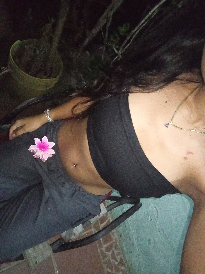 A person with long, dark hair is reclining outdoors, wearing a black strapless top and gray pants. A pink flower is placed on the left side of their abdomen, near a navel piercing. The individual is adorned with a bracelet and a necklace. A pot containing plants is visible in the background, as is a portion of a chair. The setting appears to be nighttime or dimly lit.