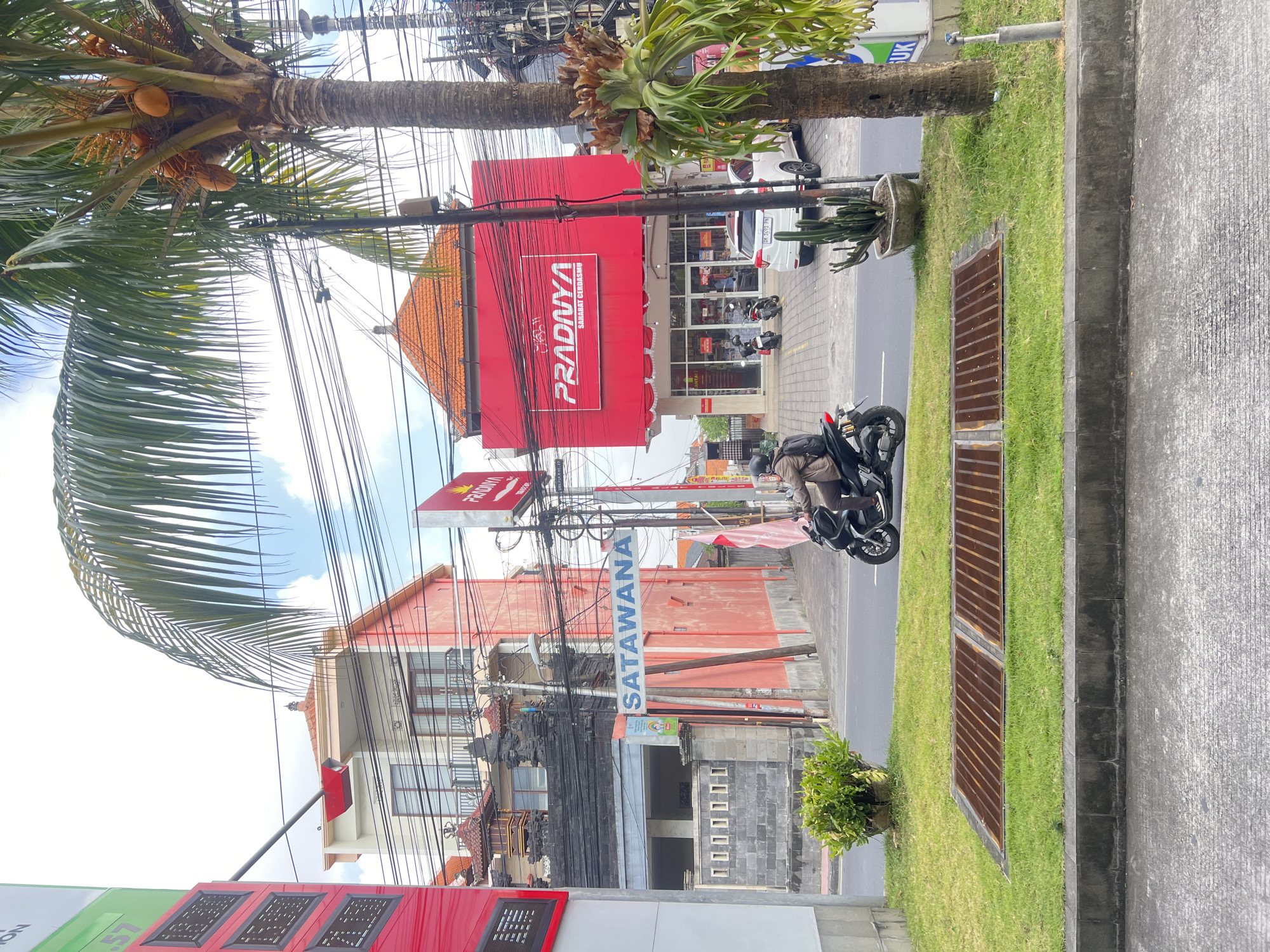 This image captures a street scene in Denpasar, Indonesia, likely on a bright, partly cloudy day. A man is riding a motorcycle on the road, heading towards the viewer's right. He is wearing a brown jacket and a helmet. In the background, a bright red building with a large sign reading "PRADNYA" is prominently visible, appearing to be a retail establishment. Next to it, another building is painted pink, and further left, a building with traditional Balinese architectural elements is visible. Overhead, a dense network of power lines crisscrosses the sky, a common sight in many Indonesian cities. A palm tree with coconuts stands to the right, adding to the tropical ambiance. A white car is parked in front of the "PRADNYA" building. The foreground shows a grassy median with a metal grate, separating the road from the sidewalk and buildings. The scene conveys a sense of everyday life in a bustling urban environment in Indonesia.