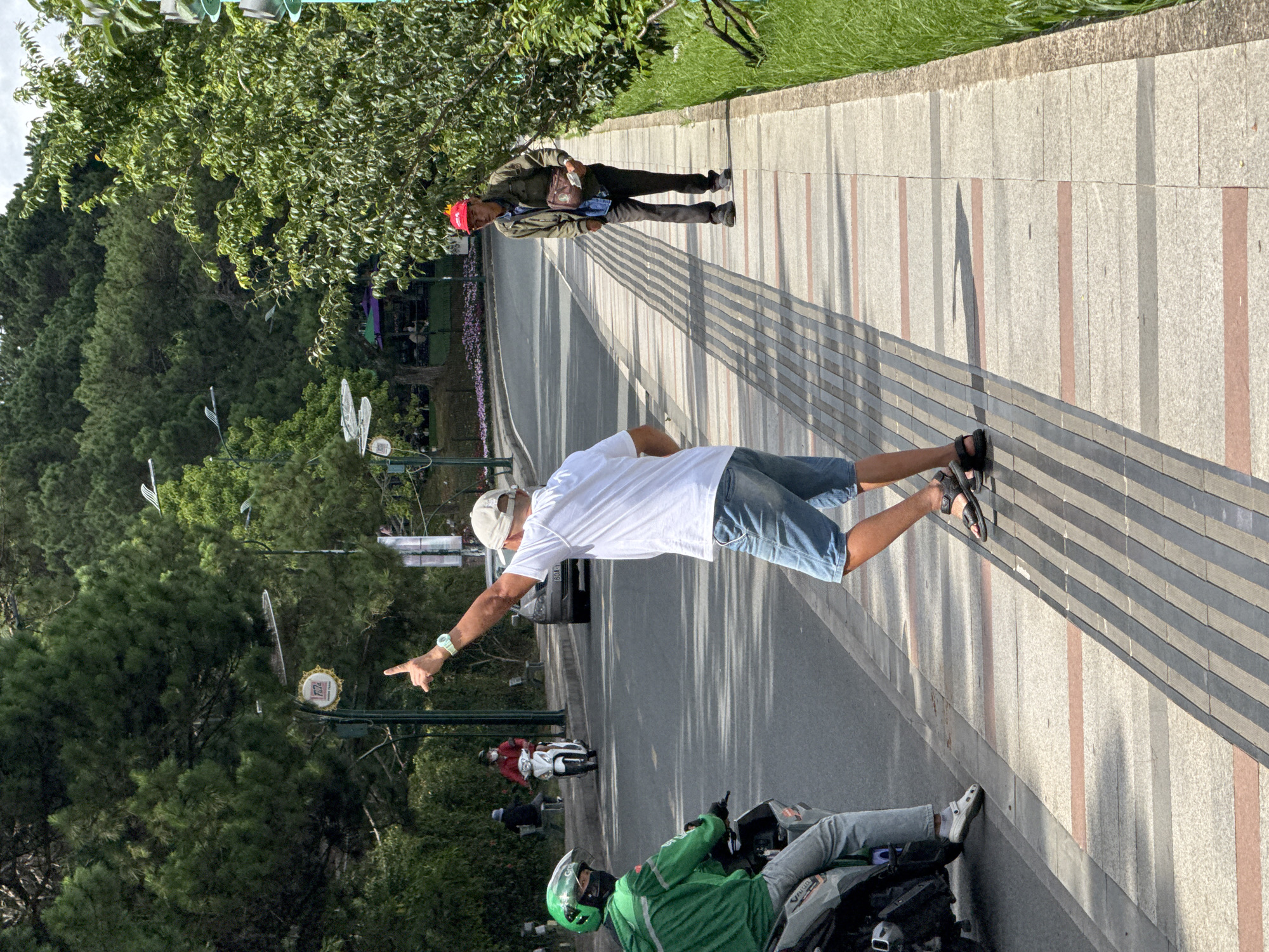 The image shows a street scene in Da Lat, Vietnam. In the foreground, a man wearing a white t-shirt, denim shorts, and sandals is standing on a sidewalk and pointing upwards with his right hand. He is facing away from the camera. To his left and slightly in front, a person wearing a green jacket and a green helmet is seated on a scooter, also facing away from the camera. Further down the street, a man in a red cap, olive green jacket, and dark pants is walking on the sidewalk. In the background, there is a road with some traffic, including a scooter with a rider wearing red and a car. The environment consists of lush green trees and foliage lining the street and sidewalk, with a cloudy sky above. The sidewalk is paved with stone tiles.