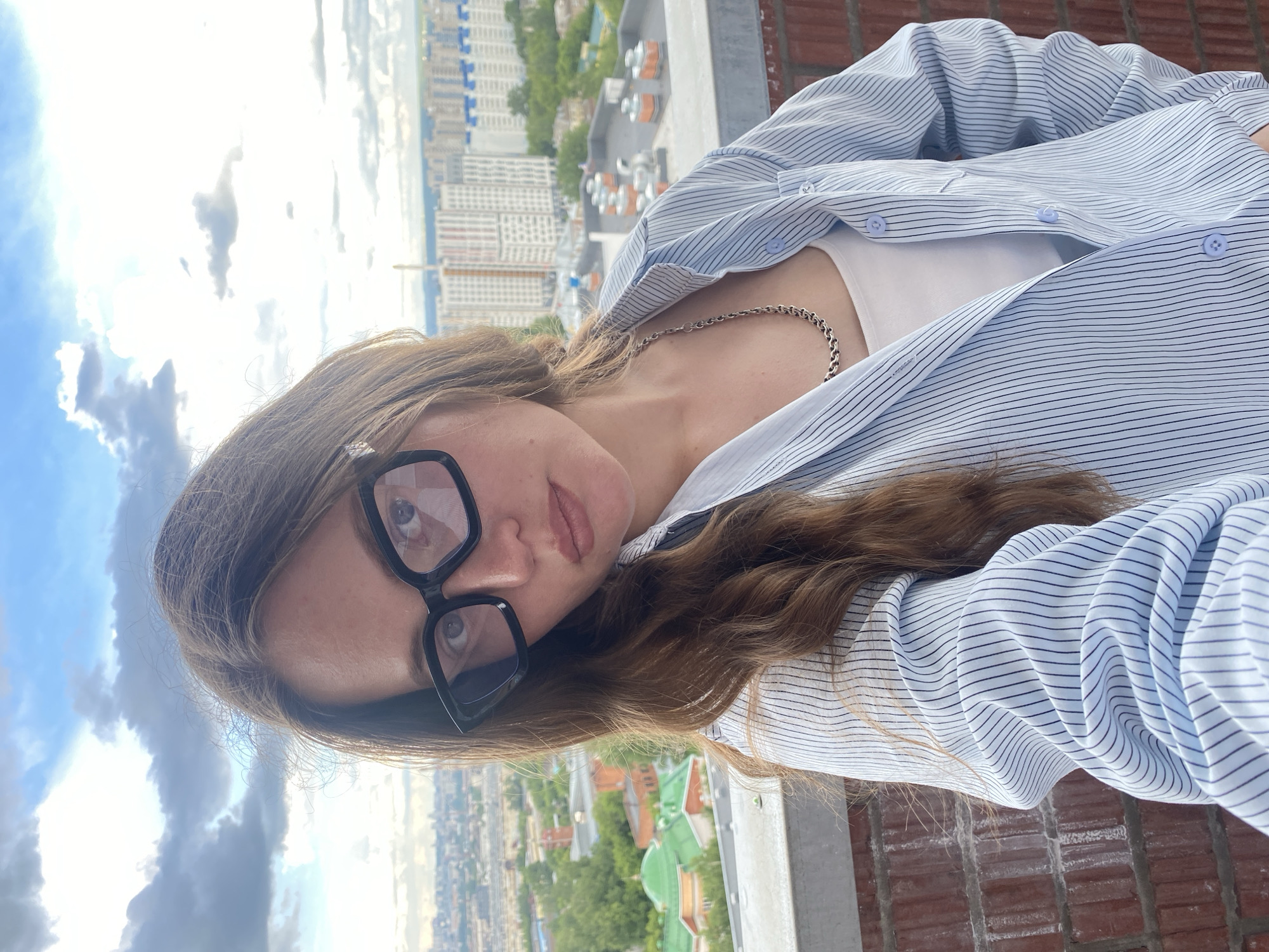 A woman with brown hair and glasses poses on a balcony. She wears a striped, button-down shirt over a white top, with a silver chain necklace visible. The background features a city view, identifiable as Izhevsk, Russia, with a mix of high-rise apartment buildings and green spaces. The sky is partially cloudy. The balcony's edge is made of red brick.
