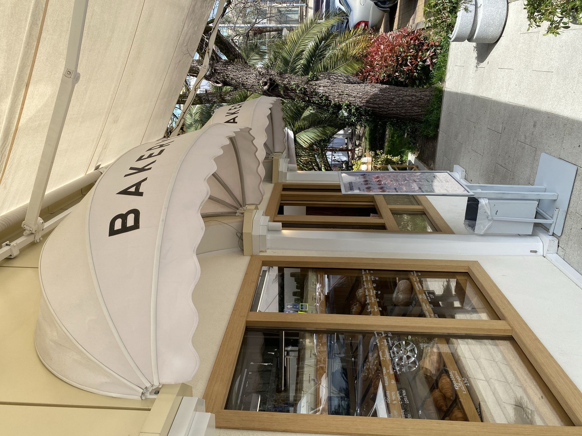 This is a photograph of a bakery storefront in Sochi, Russia. The facade of the bakery is cream-colored with wooden-framed windows displaying various baked goods. The sign on the canopy reads "BAKERY" in large, bold letters. A menu is posted at the entryway. Beyond the bakery, a palm tree and some greenery are visible, along with a glimpse of a silver car and a park. The scene is bathed in daylight.