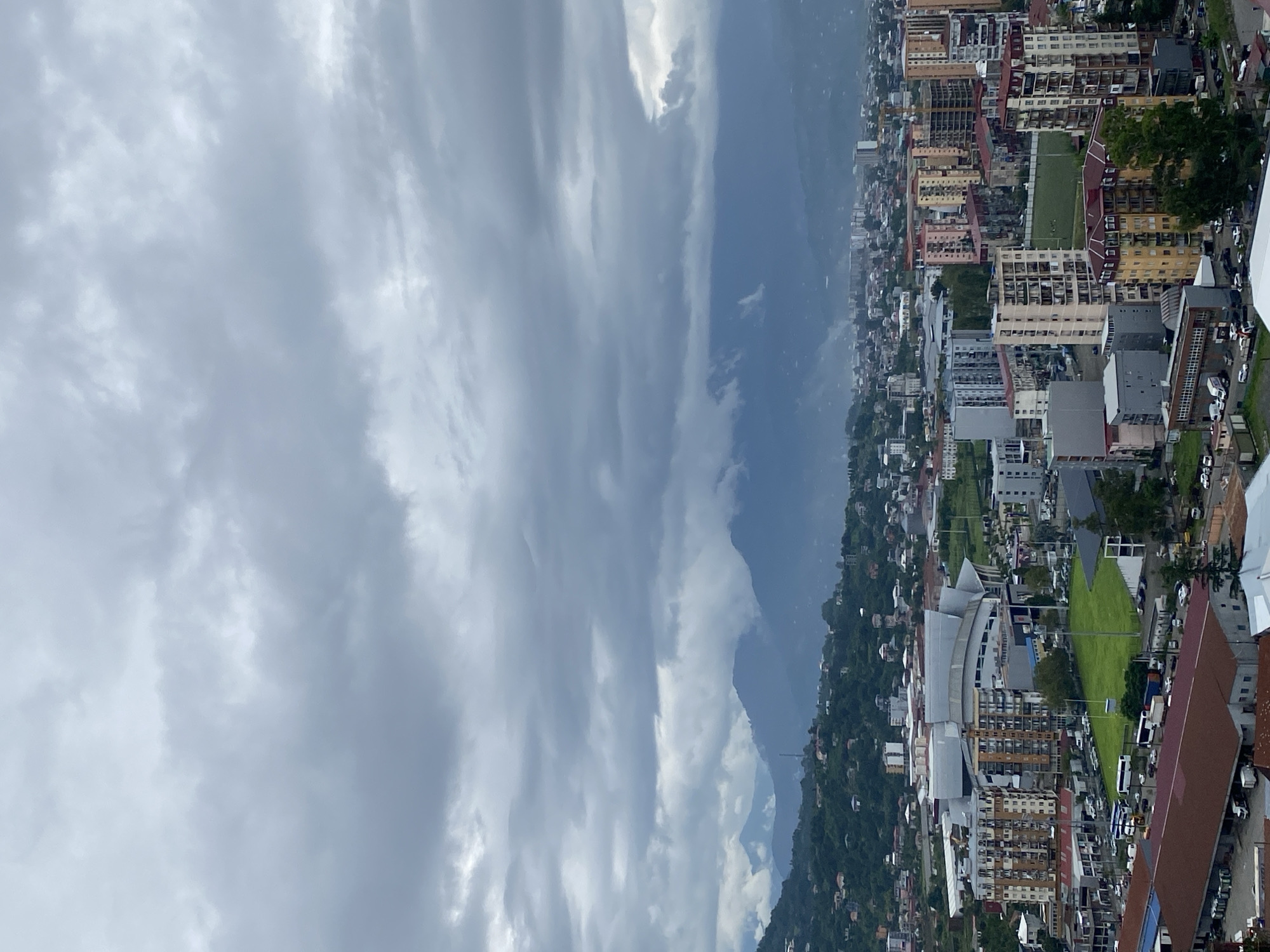 The image captures a panoramic view of Batumi, Georgia, on a cloudy day. In the foreground, there are several multi-story buildings, including residential complexes and commercial structures. Some buildings feature colorful facades and balconies, while others are more utilitarian in design. A large sports complex with a distinctive roof structure is visible in the mid-ground, surrounded by parking areas and green spaces. Beyond the urban landscape, rolling hills covered in lush vegetation lead to majestic mountains in the distance, shrouded in mist and clouds. The sky is overcast with heavy, grey clouds, suggesting a potentially rainy or stormy weather condition. The overall scene depicts a developing urban environment nestled between natural landscapes.