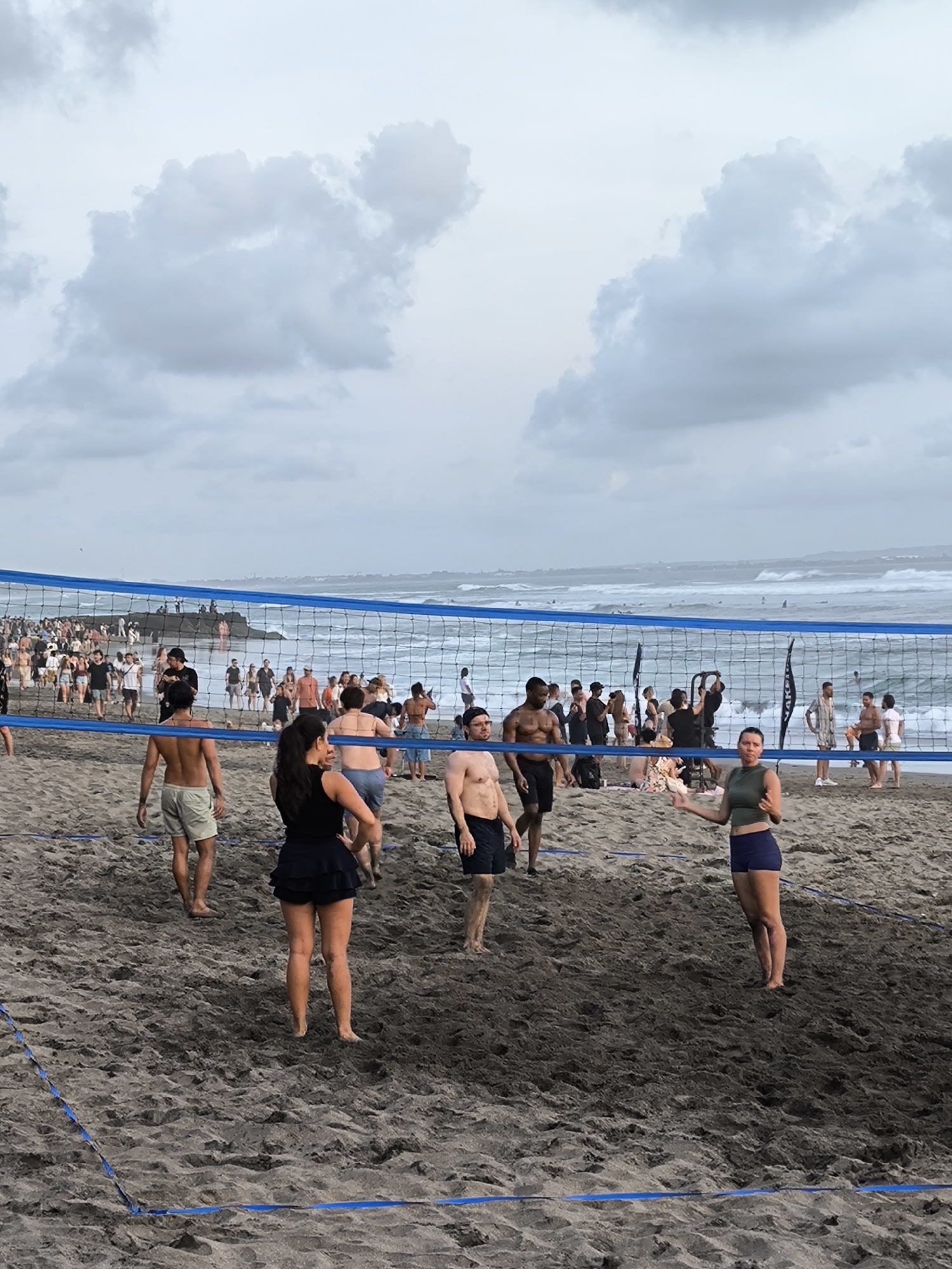 This image captures a scene at Seminyak Beach in North Kuta, Indonesia, on a cloudy day. The foreground features a sandy volleyball court with a blue net dividing the playing area. Several individuals are visible on the court, some in athletic attire, and others in beachwear, suggesting a casual game or social gathering.
In the mid-ground, a large group of people are gathered on the beach, some appearing to be spectators or participants in activities. Beyond them, the ocean stretches out, with moderate waves and a few surfers or paddleboarders in the water. The horizon is visible under a sky filled with dramatic, grey clouds. The overall atmosphere suggests a lively beach environment with recreational activities taking place.