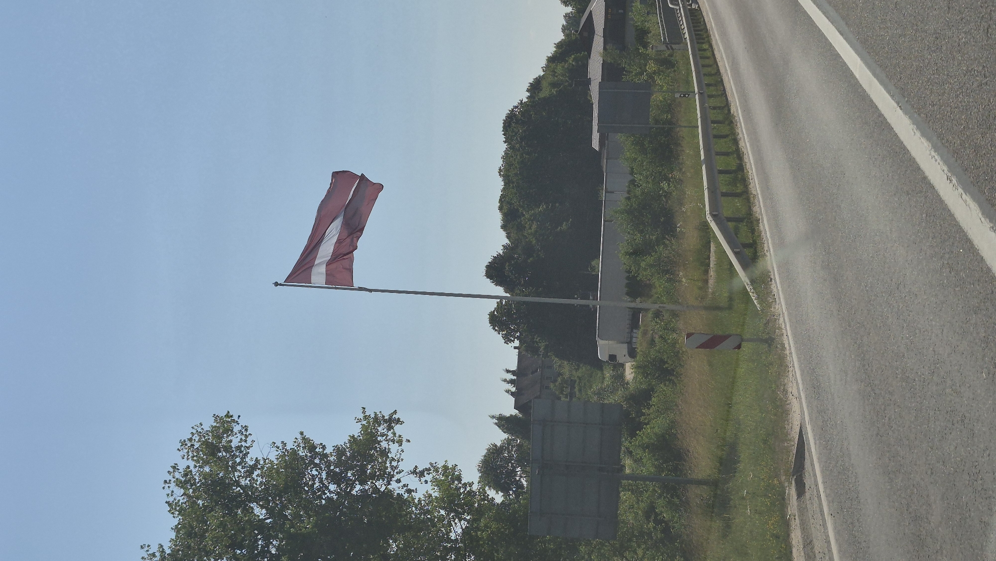 The image depicts a roadside scene under a clear blue sky. A Latvian flag flies on a flagpole to the right of center. In the background, there is a dense line of trees, and beyond that, some buildings are visible. A white truck is parked on the side of the road near the trees. A metal guardrail runs along the edge of the road in the foreground. The road itself is paved and appears to be a highway or major road. The overall environment suggests a rural or semi-rural setting. The location is the city of Smėlynė, Lithuania.
