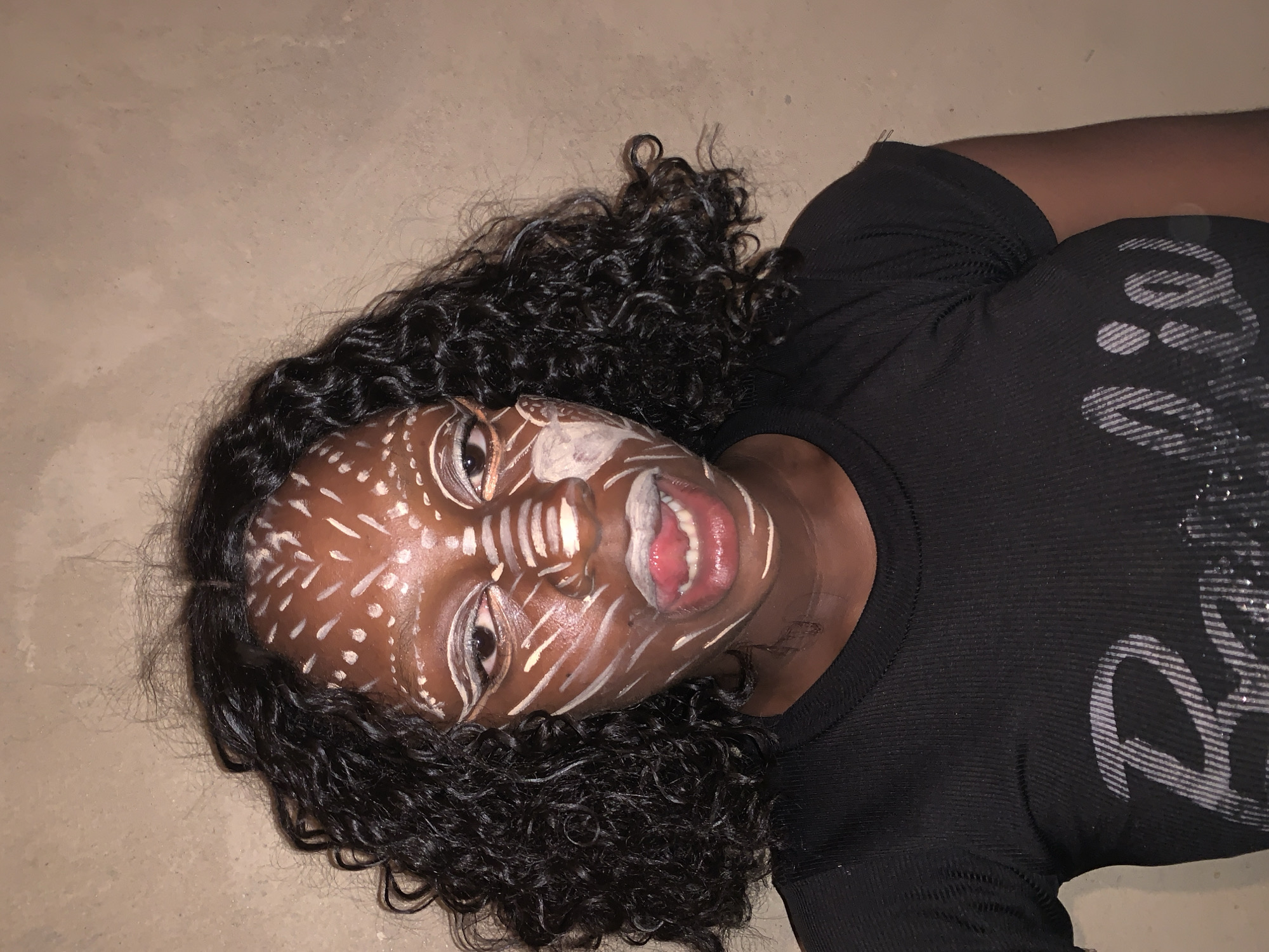 A dark-skinned person with curly black hair lies on a light brown surface. Their face is painted with white markings resembling tribal patterns. They wear a black, tight-fitting shirt with some sort of decoration. The background is plain and featureless. The location in the city of Agban, Nigeria, cannot be confirmed, as there are no visual cues in the image to ascertain a geographical location.
