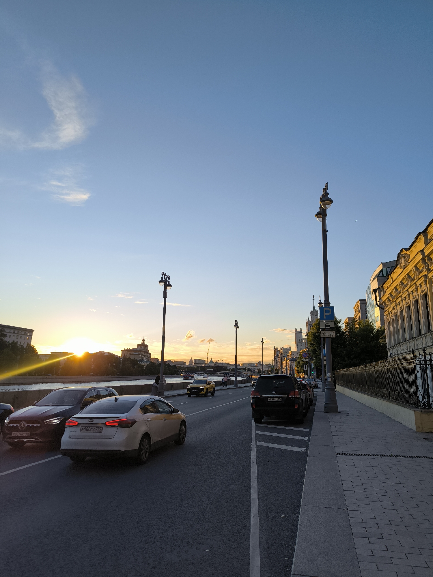 The image depicts a street scene in Moscow, Russia, during sunset. A street with cars and a sidewalk lines the right side of the frame. The sun is setting over a river and buildings on the left, casting a warm glow and lens flare. Several cars are visible on the street, including a white Kia sedan with a taxi sign, a dark Mercedes-Benz, and a black SUV. A lamppost with multiple lights stands on the sidewalk. In the background, more buildings and a bridge are visible, along with a crescent moon in the sky. The overall atmosphere is serene, with the warm colors of the sunset dominating the scene.
