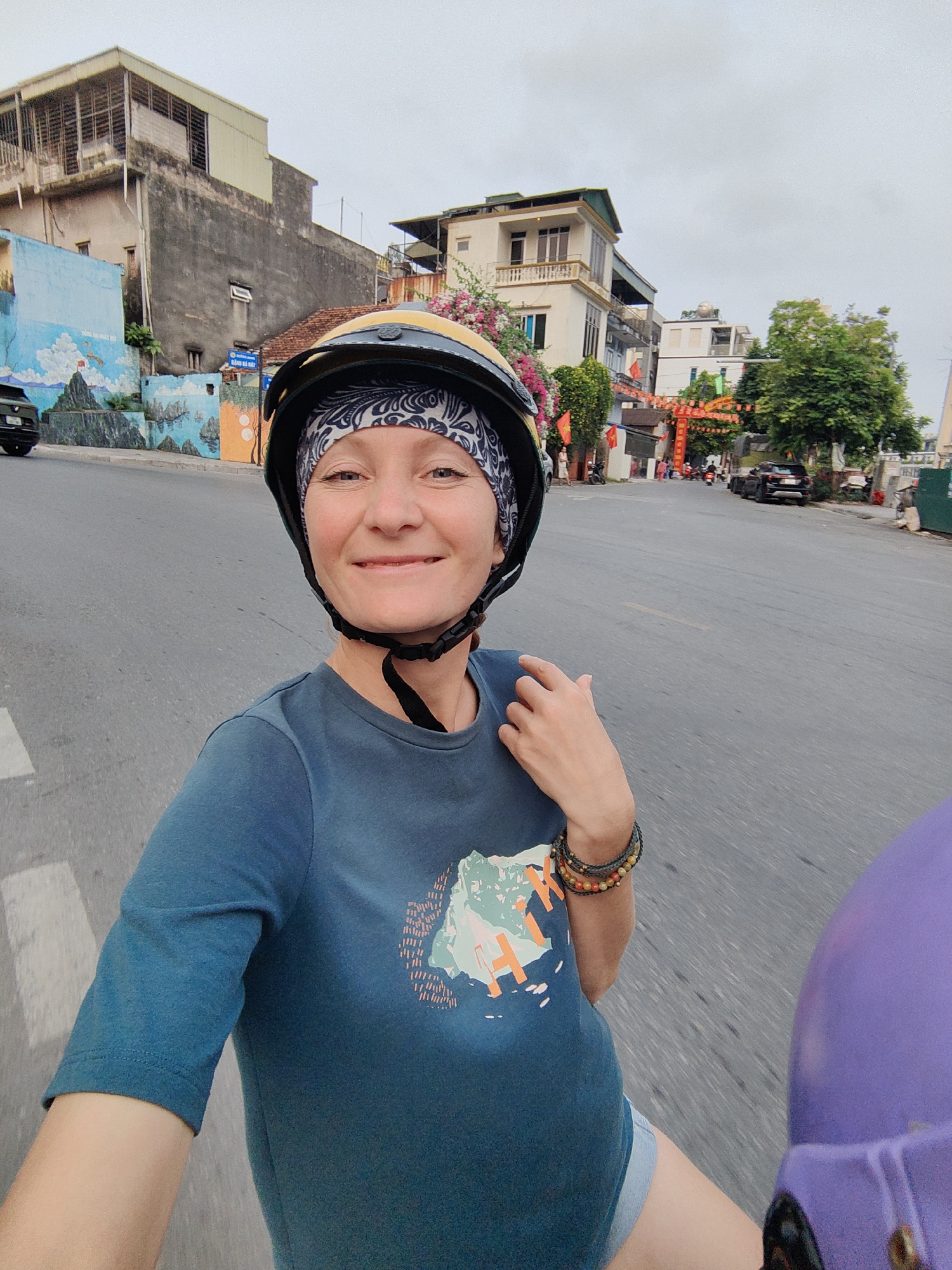 A woman is riding a motorcycle in Hong Gai Ward, Vietnam, as indicated by the painted mural on the wall showing Halong Bay and street decorations that are present. She is wearing a blue t-shirt with a nature print, a patterned bandana and a yellow and black helmet. She has bracelets on her left wrist. She is smiling at the camera. The roadway has a center line. Buildings, decorative archway, parked cars and trees line the roadway.