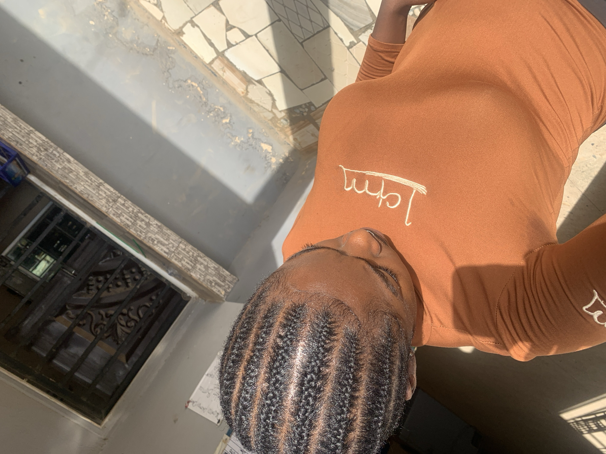 A medium shot of a person indoors near New Stadium, Nigeria. The person is wearing a brown long-sleeved top with white lettering and has cornrows. They are standing near a window with bars and a mosaic-tiled floor. The floor is light-colored and the wall is grey. The lighting appears to be a mix of natural and artificial.