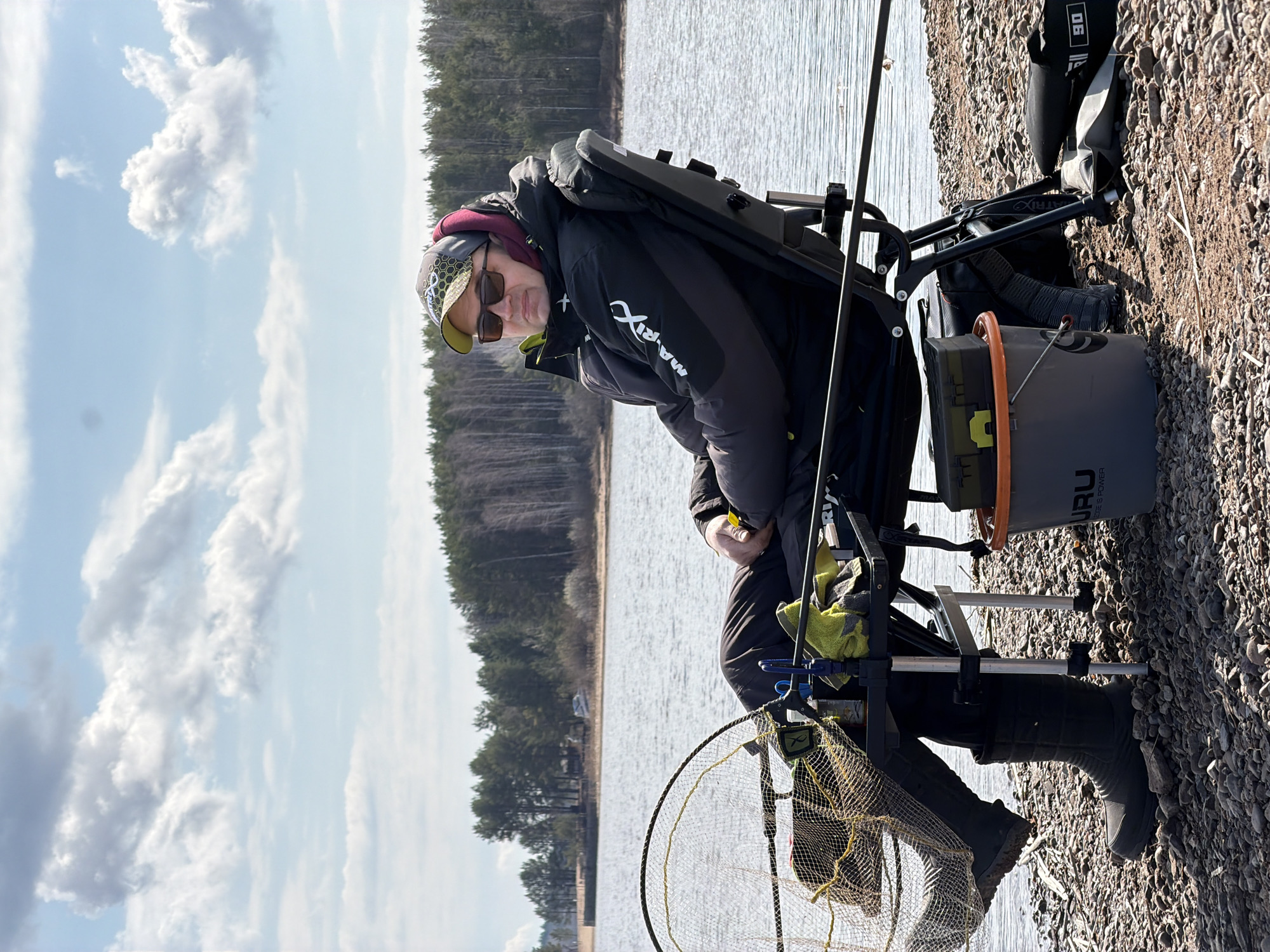 The image depicts an individual seated in a fishing chair near a body of water. The person is wearing a dark jacket, sunglasses, and a hat. Fishing gear, including a net and a bucket labeled "GURU," surrounds the chair. The setting appears to be a rocky shoreline with a forest visible in the background. The sky is partly cloudy. Based on the prompt, this location is likely near the city of Chaikovsky, Russia. The individual seems to be engaged in the activity of fishing.