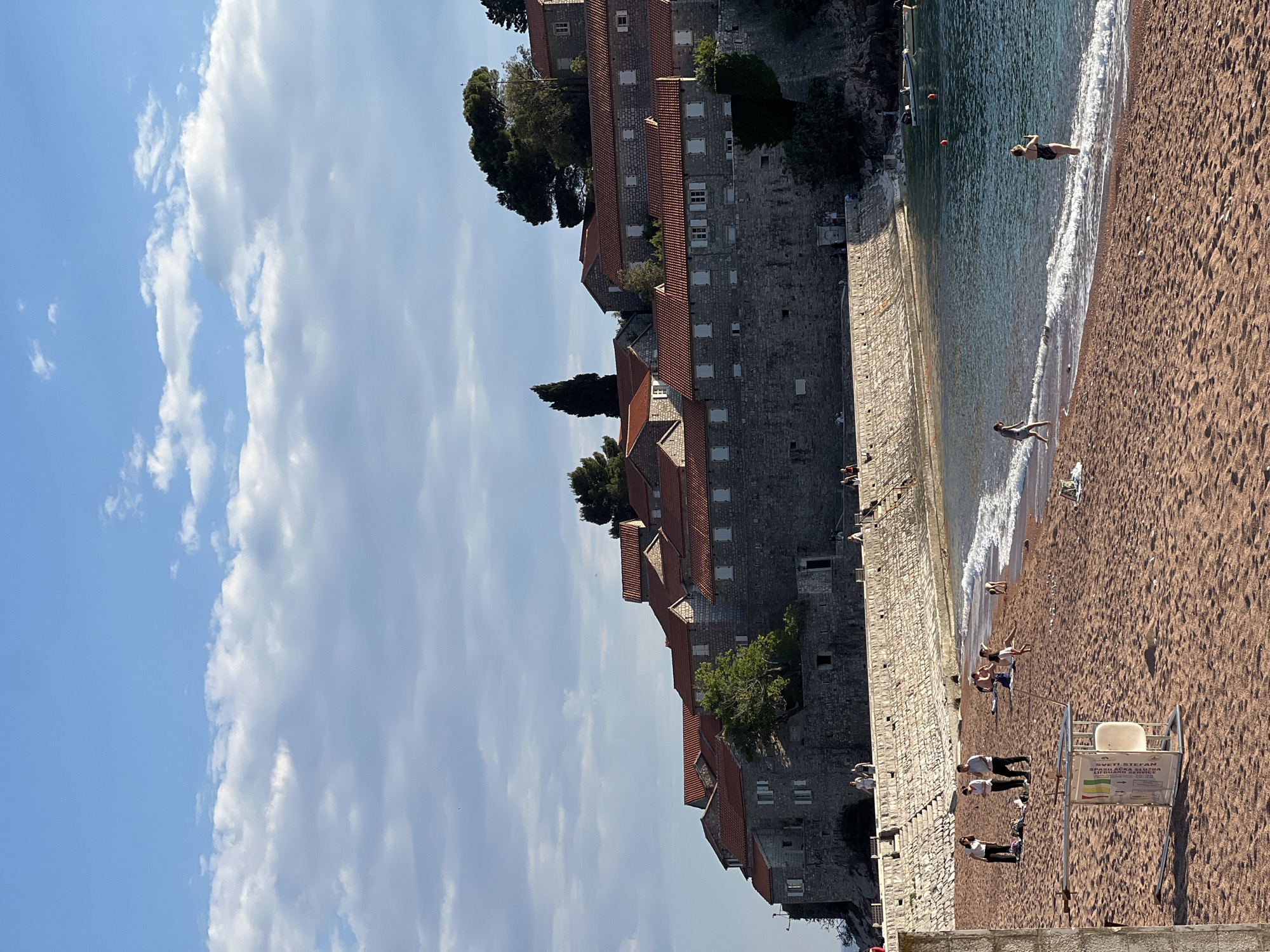 The image shows a beach scene in Sveti Stefan, Montenegro, with a historic walled town to the side of the beach. The beach is a reddish-brown color, and the sea is turquoise. People are swimming and sunbathing along the shore. A lifeguard stand is on the beach. The sky is light blue with white clouds.