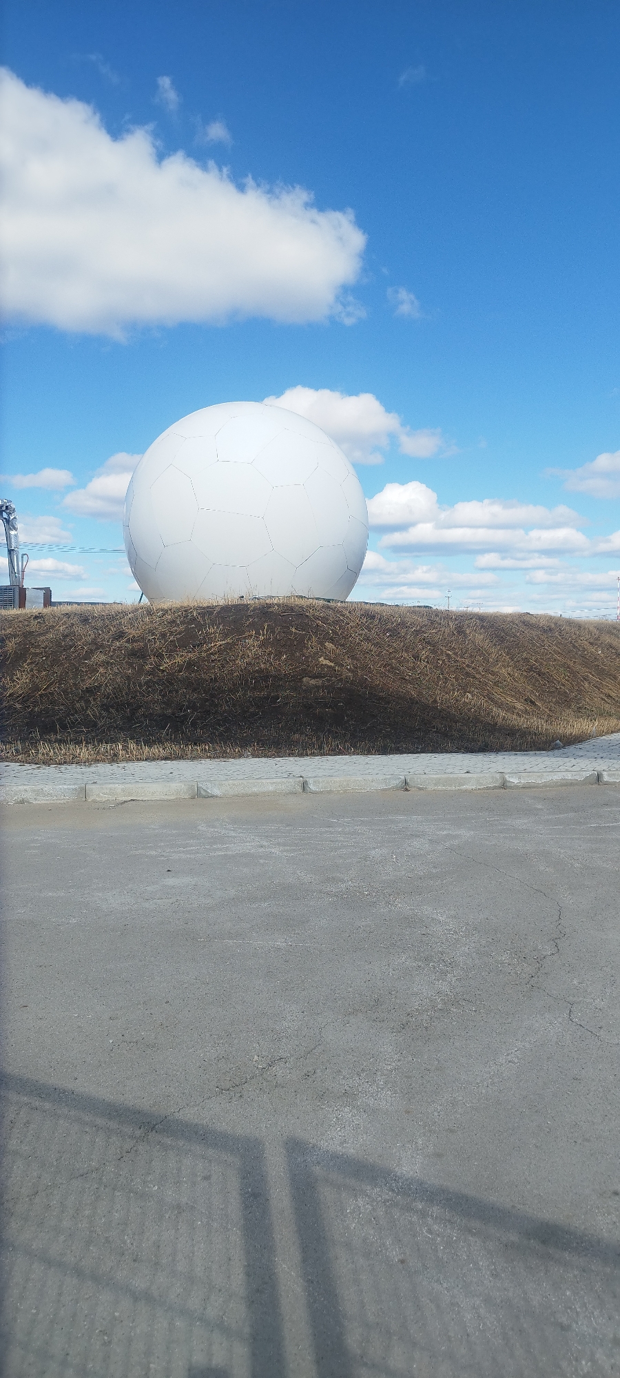 The image shows a radome located in Yakutsk, Russia. The radome is a large, white, spherical structure built on top of a small hill covered in dry grass. It is segmented with a pattern of interconnected polygons. The sky is blue with scattered white clouds. In the foreground, there is a paved asphalt surface with a concrete curb separating it from the hill. The shadow of a fence or other structure is cast on the asphalt.