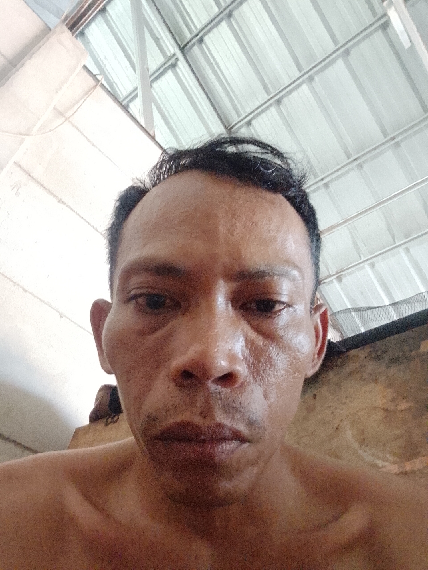 A close-up, low-angle headshot of a man with dark hair and complexion, under a corrugated metal roof structure. The man has short, dark hair with some grey strands. He has a tired expression. The environment is inside a building in what appears to be Bukit Ketok, Indonesia.