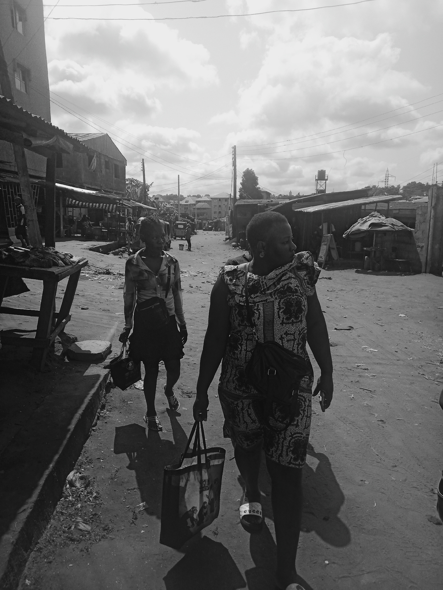 Two women are walking down a dusty street in Onitsha, Nigeria. The woman in the foreground is wearing a patterned dress and carrying a shopping bag. The other woman is wearing a collared shirt and a short skirt. They are both carrying shoulder bags. The street is lined with makeshift shops and stalls, and there is litter on the ground. The sky is cloudy.