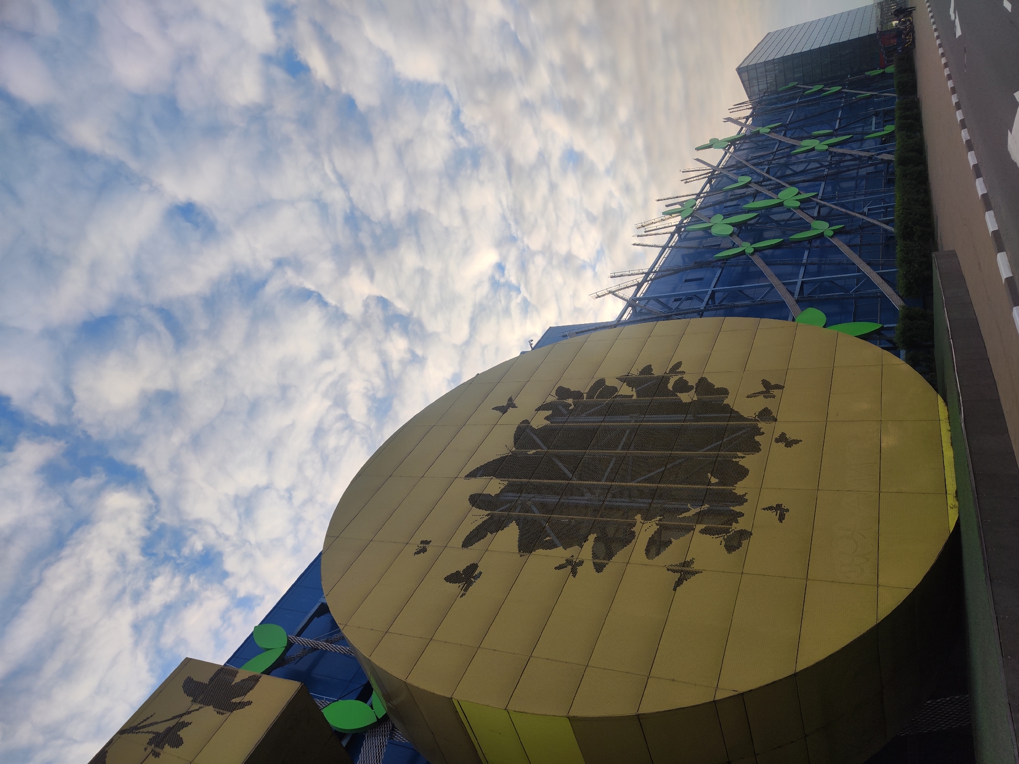 A striking low-angle view captures a unique, modern building under a partly cloudy sky in Saint Petersburg, Russia. The most prominent feature is a large, golden, circular facade that dominates the foreground and mid-ground. This golden surface is perforated with numerous dark cutouts forming a detailed pattern of butterflies, some scattered individually and others densely clustered.
Rising behind and above this golden element is the main structure of the building, characterized by a reflective blue glass facade. Adorning this blue section is a diagonal lattice-work from which large, bright green, stylized leaf-shaped structures extend outwards, adding a naturalistic and decorative touch. To the lower left, another smaller, rectangular golden panel is visible, also featuring a dark, leaf-like cutout design.
Further up, the building appears to continue with more conventional glass-panelled sections. On the far right, a segment of a road or pathway is visible, edged by a curb and marked with white and black dashed lines. A narrow strip of manicured green bushes runs along the building's base beside this path.
The sky is bright blue with scattered, wispy white clouds, indicating daytime. The light is diffused, suggesting either early morning or late afternoon rather than harsh midday sun. The overall impression is one of innovative and artistic architecture, blending modern materials with organic themes. There is no visible text in the image.