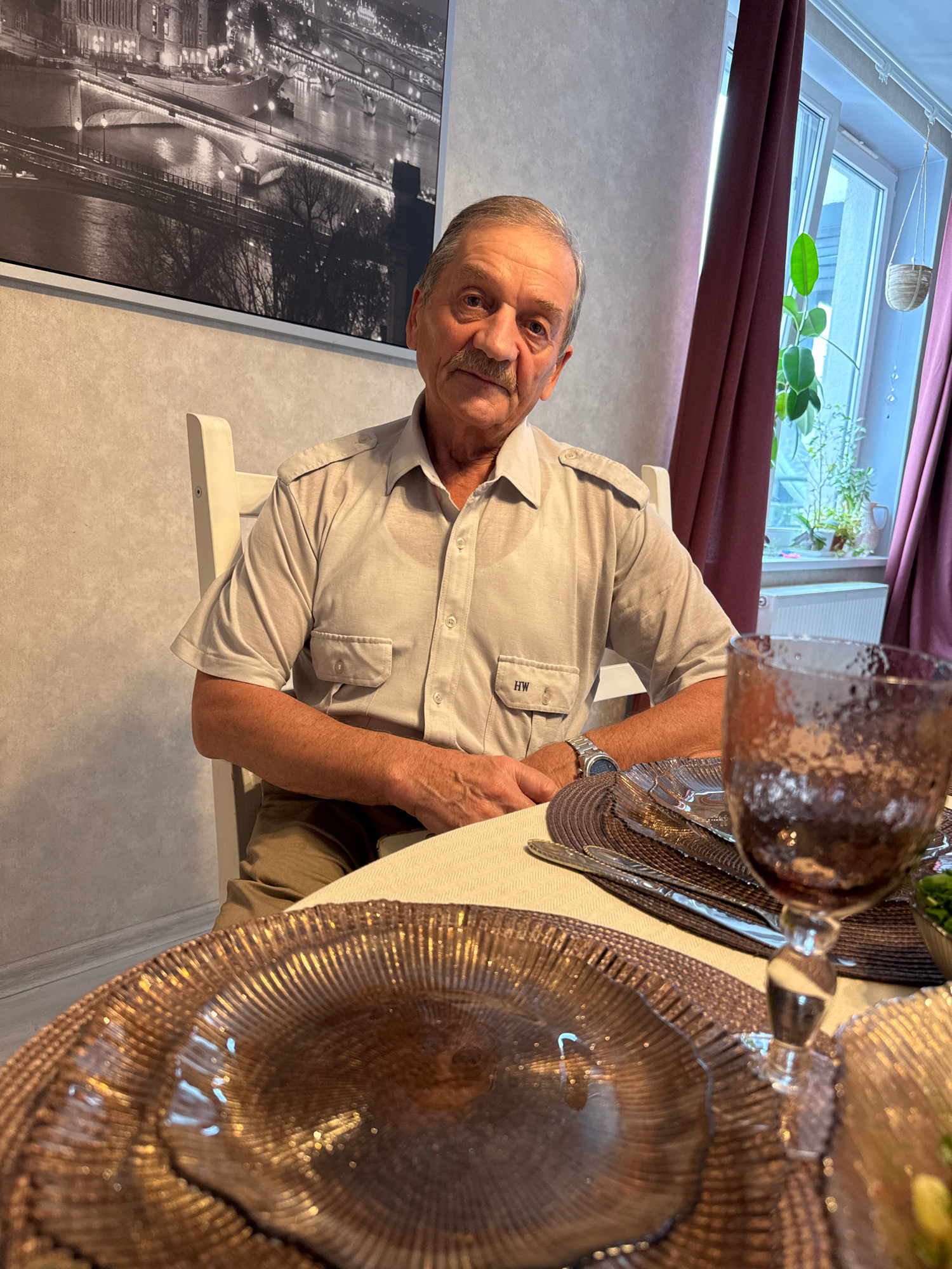An elderly man with a mustache, wearing a light-colored short-sleeved button-down shirt and khaki pants, sits at a dining table set for a meal. He is seated in a white wooden chair, with his hands resting in his lap. The table is covered with a textured tablecloth, and place settings include metallic chargers, plates, and cutlery. A glass filled with a dark liquid is visible in the foreground. Behind the man, a large black and white photograph of a cityscape, possibly Paris, hangs on the wall. To the right, a window is draped with dark purple curtains, and several potted plants are visible on the windowsill. The overall scene appears to be an indoor dining setting, likely a home. The mention of Saint Petersburg, Russia, in the prompt seems unrelated to the visual content of the image.