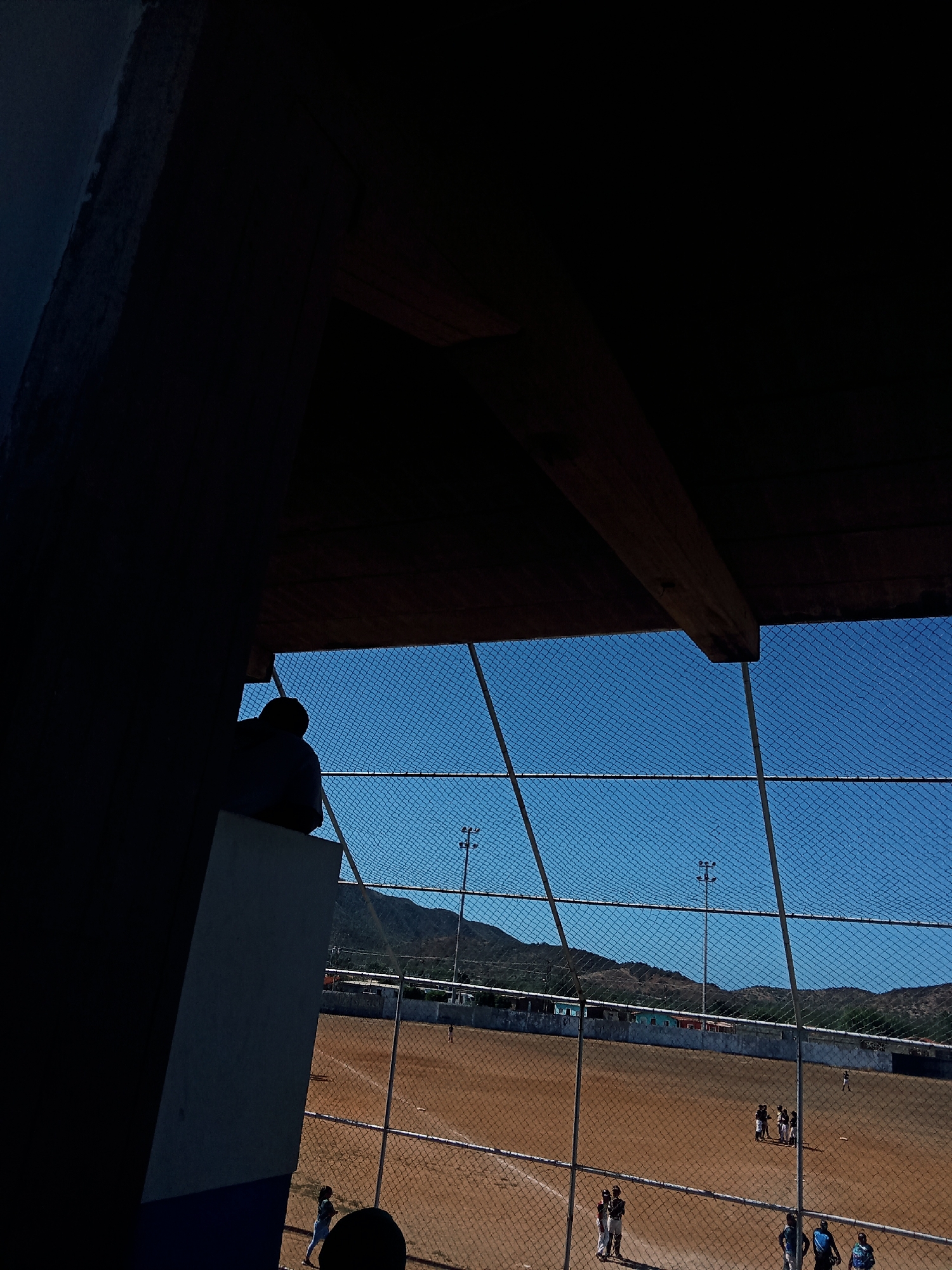 The image shows a baseball field in Juan Griego, Venezuela, viewed through a chain-link fence. The field is dirt, and several people are on it, including some who appear to be players in uniform. A mountain range is visible in the background behind a low, concrete wall bordering the field. Two utility poles stand in the distance. The photo is taken from beneath a wooden shelter structure, obscuring the upper left corner of the view.