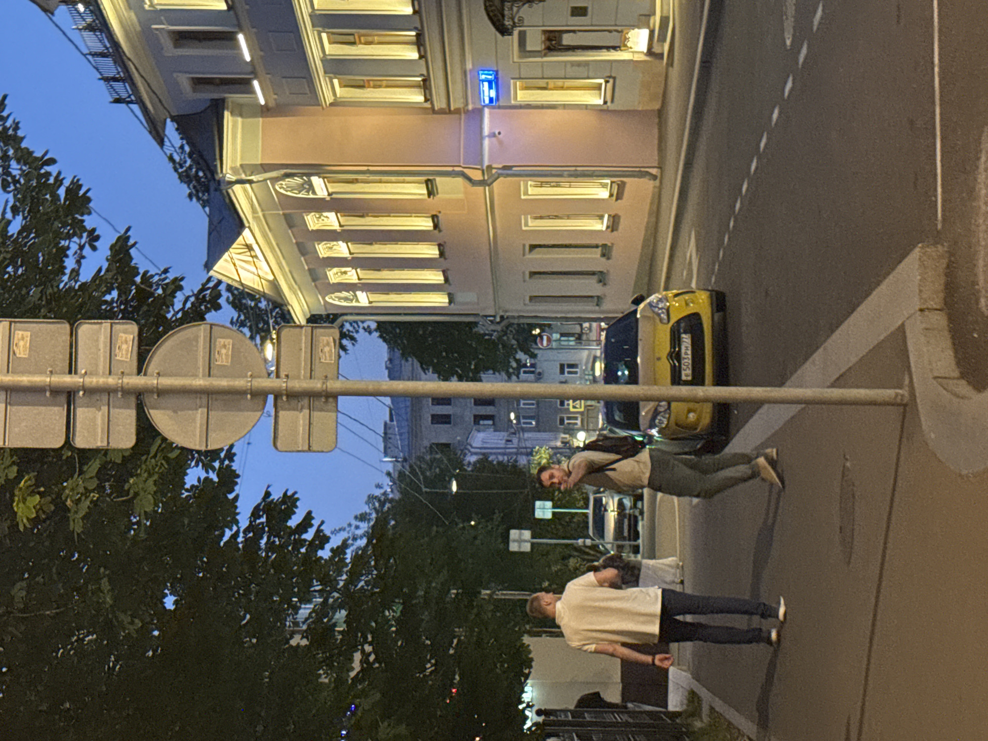 The image captures a street scene in Moscow, Russia, at dusk or early evening. Two men are walking on a sidewalk on the left side of the frame. The man closer to the foreground is wearing a light-colored polo shirt and dark pants, with his left arm extended downwards. The man further back, on the right, is wearing a light brown t-shirt and grey pants and is looking towards the camera, pointing with his right index finger. He has a backpack slung over his left shoulder.
In the middle of the street, a yellow Citroën car is parked. The car has a license plate visible which appears to read "E503PH 77". Behind the car, other vehicles and buildings can be seen. The buildings on the right side of the street are lit from within, with multiple windows illuminated, and are painted in pastel colors, including pink and light blue. A blue street sign with the number "7" is affixed to one of the buildings.
A tall pole with multiple traffic or informational signs is positioned in the foreground, slightly off-center to the right. Trees with dark green foliage line the left side of the street, with some leaves partially obscuring the sky. The sky is a deep blue, indicating the transition from day to night. The road surface is asphalt, marked with dashed white lines. The sidewalk is paved and bordered by a concrete curb.