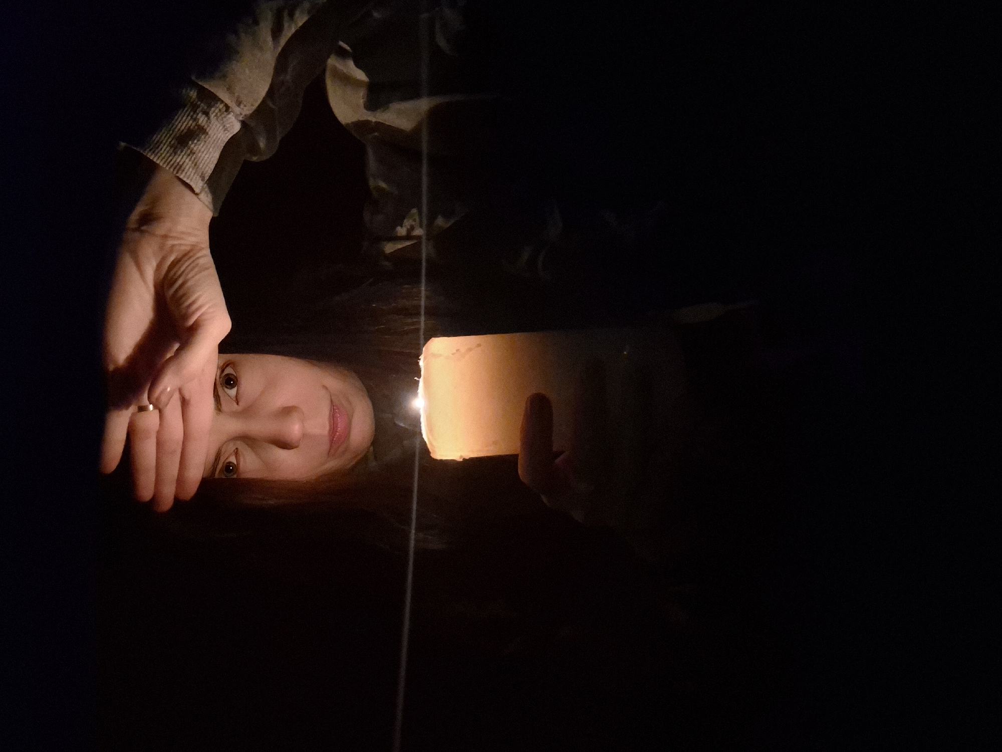 The image displays a person's upper body, oriented sideways with their head to the left, illuminated by a candle in a predominantly dark setting. The subject is a female with dark hair; her face is partially visible on the left side of the frame, lit by a large, white pillar candle held in her right hand (from her perspective). Her bright eyes gaze directly forward, and a faint, gentle smile graces her lips. Her left hand is raised, resting on her forehead, revealing a ring on her ring finger and the cuff of a long-sleeved garment. The candle serves as the primary light source, casting a warm glow on her skin and the surrounding area. The background is almost entirely black, indicating extremely low ambient light, suggesting the scene takes place at night or in a very dark room. A faint, thin vertical line runs through the center of the image, possibly a reflection or a structural element. The scene captures a moment of quiet contemplation or intimacy. No text is visible in the image. The provided location context is Chaikovsky, Russia.
