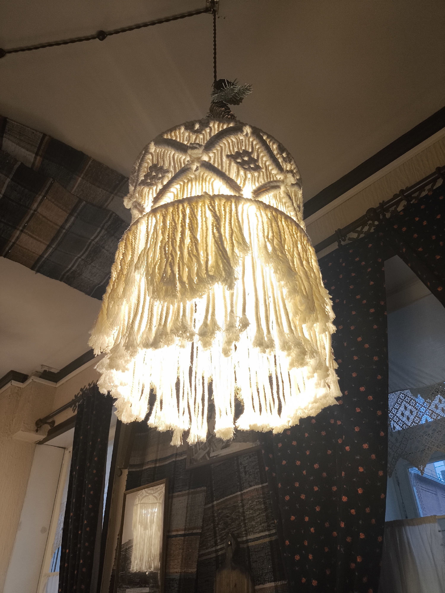 A macrame chandelier with two tiers of fringe hangs from the ceiling. The chandelier is lit from within, casting a warm glow. In the background, there are dark curtains with a floral pattern and a plaid-patterned wall hanging. The scene appears to be an interior shot, possibly in a residential or commercial space. There is no information available about the specific location in Perm, Russia.
