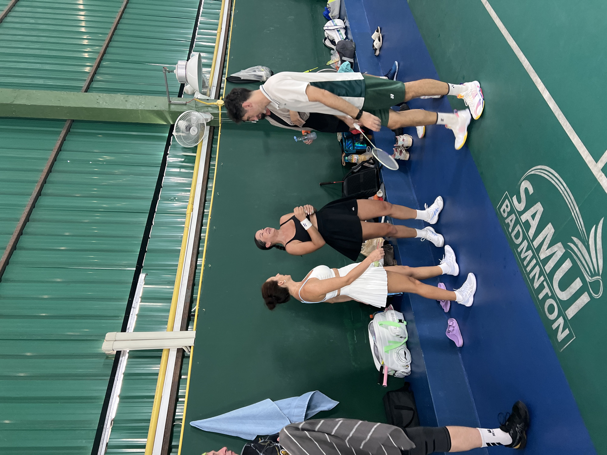 In a badminton court in Ban Bang Rak, Thailand, a group of four people are gathered. Two women, dressed in athletic wear consisting of white and black tennis skirts and tops, are laughing. One woman is wearing a white pleated skirt and a white sleeveless top, while the other is in a black short skirt and a black tank top. A man in a white polo shirt and dark green shorts is holding a badminton racket. Another person, partially visible on the left, is wearing athletic shorts, knee-high socks, and a black t-shirt with a striped pattern. The court features a blue and green floor with white markings. Bags, water bottles, and shoes are scattered on a bench along the wall. The background consists of a green wall and a corrugated metal ceiling.