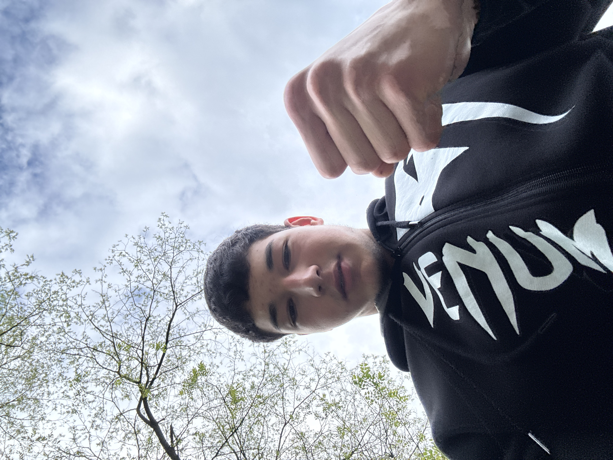 A low-angle shot shows a young man looking down at the camera. He has short dark hair and is wearing a black hoodie with white lettering. A hand is clenched into a fist above his head. The background includes trees with budding leaves and a cloudy sky. The location in the city of Zlatograd, Bulgaria is not discernible from the image.