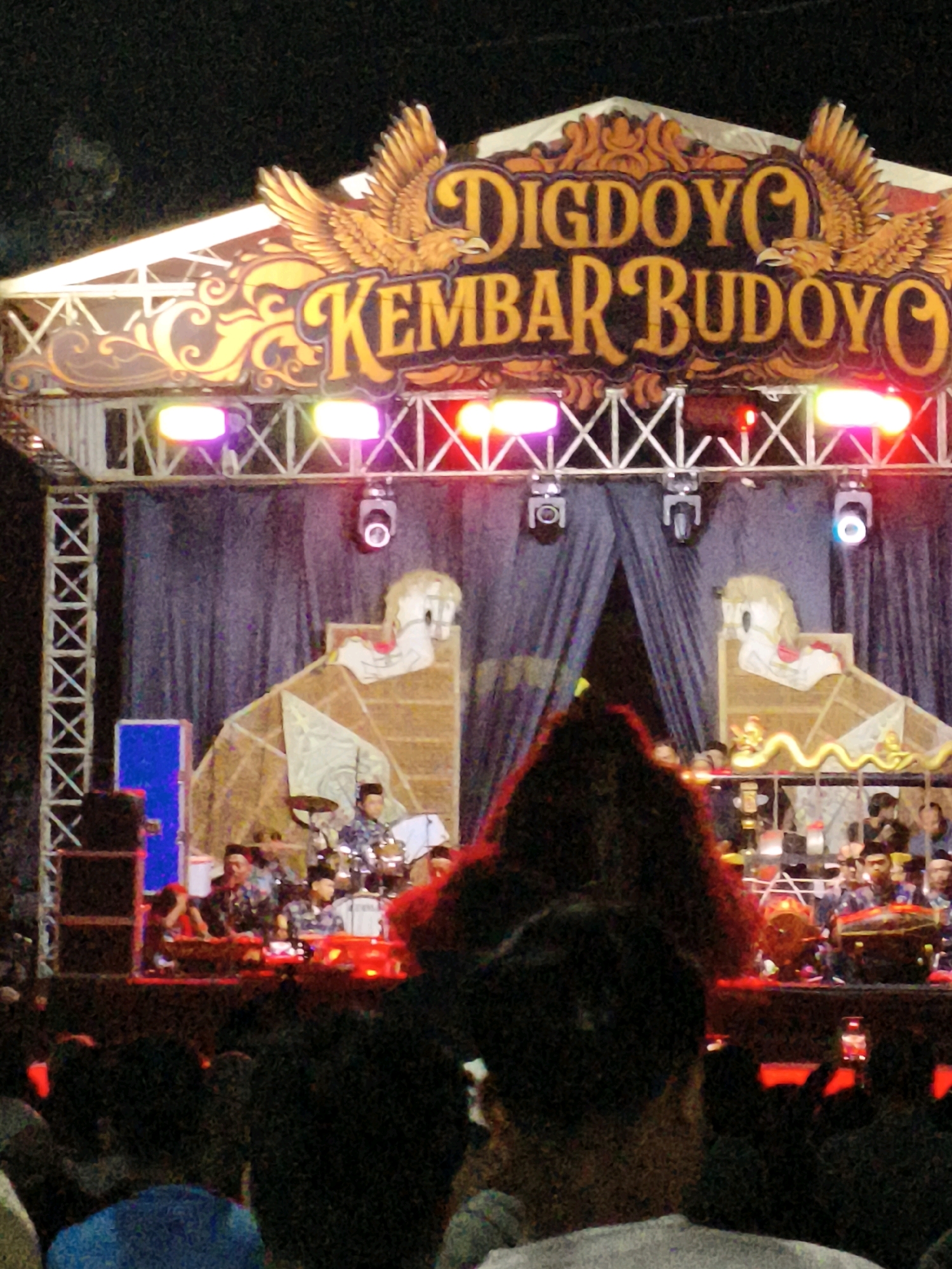 The image depicts an outdoor stage at night, likely for a cultural performance or festival, in Kembaran, Indonesia. The stage is adorned with a large, ornate sign featuring the words "DIGDOYO KEMBAR BUDOYO" in stylized lettering, accompanied by decorative golden eagles on either side. Below the sign, a colorful lighting rig illuminates the stage with red, pink, and purple lights.
The stage itself has a dark blue curtain backdrop. In the center, there are two large, elaborately decorated structures resembling stylized horses or mythical creatures, possibly part of a parade float or stage decoration. A group of musicians is visible on stage, some playing drums and other percussion instruments. They are wearing traditional clothing.
In front of the stage, an audience is gathered, with the foreground dominated by the backs of several audience members. The atmosphere appears to be festive and lively, suggested by the stage setup and the presence of a crowd. The lighting suggests it is evening or nighttime.