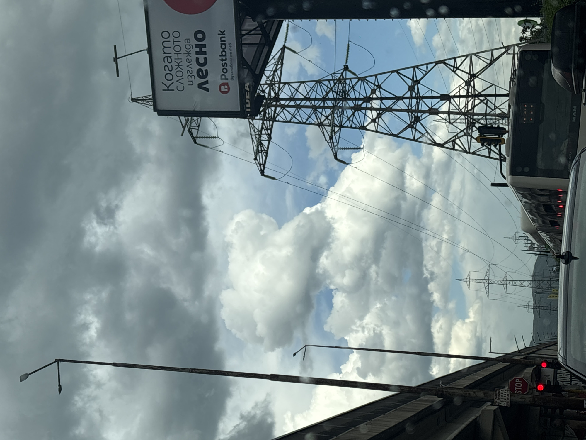 The image shows a view from a vehicle in Sofia, Bulgaria, looking up at power lines and a billboard. The billboard features advertising for Postbank and Idea. The scene includes a gray, cloudy sky with patches of blue, power line towers, a bus, and a stop sign with a traffic light. A mountain range can be seen in the distance. A sign with Bulgarian text and numbers is also visible. The photo is taken at an angle, indicating the vantage point from inside the vehicle. The road is wet, suggesting recent or ongoing rain.