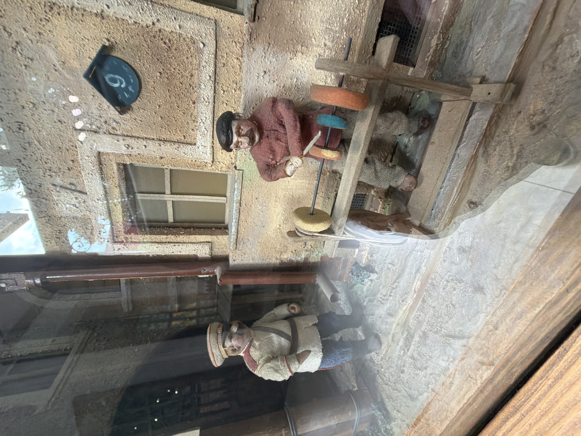 This is a diorama featuring two miniature figures in a narrow, cobblestone alleyway. The figure on the left, dressed in a military-style uniform with a peaked cap, stands with his hands at his sides. The figure on the right, wearing a red shirt and brown trousers, is engaged in an activity. He is operating a wooden contraption with a metal rod and several colorful discs, possibly a spinning wheel or a similar craft. The backdrop depicts a textured building wall with a window and a building number "9" affixed to it. The scene evokes the ambiance of an old European city, and based on the building number and architectural style, it is likely a representation of an alleyway in the Sololaki district of Tbilisi, Georgia, known for its historic charm.