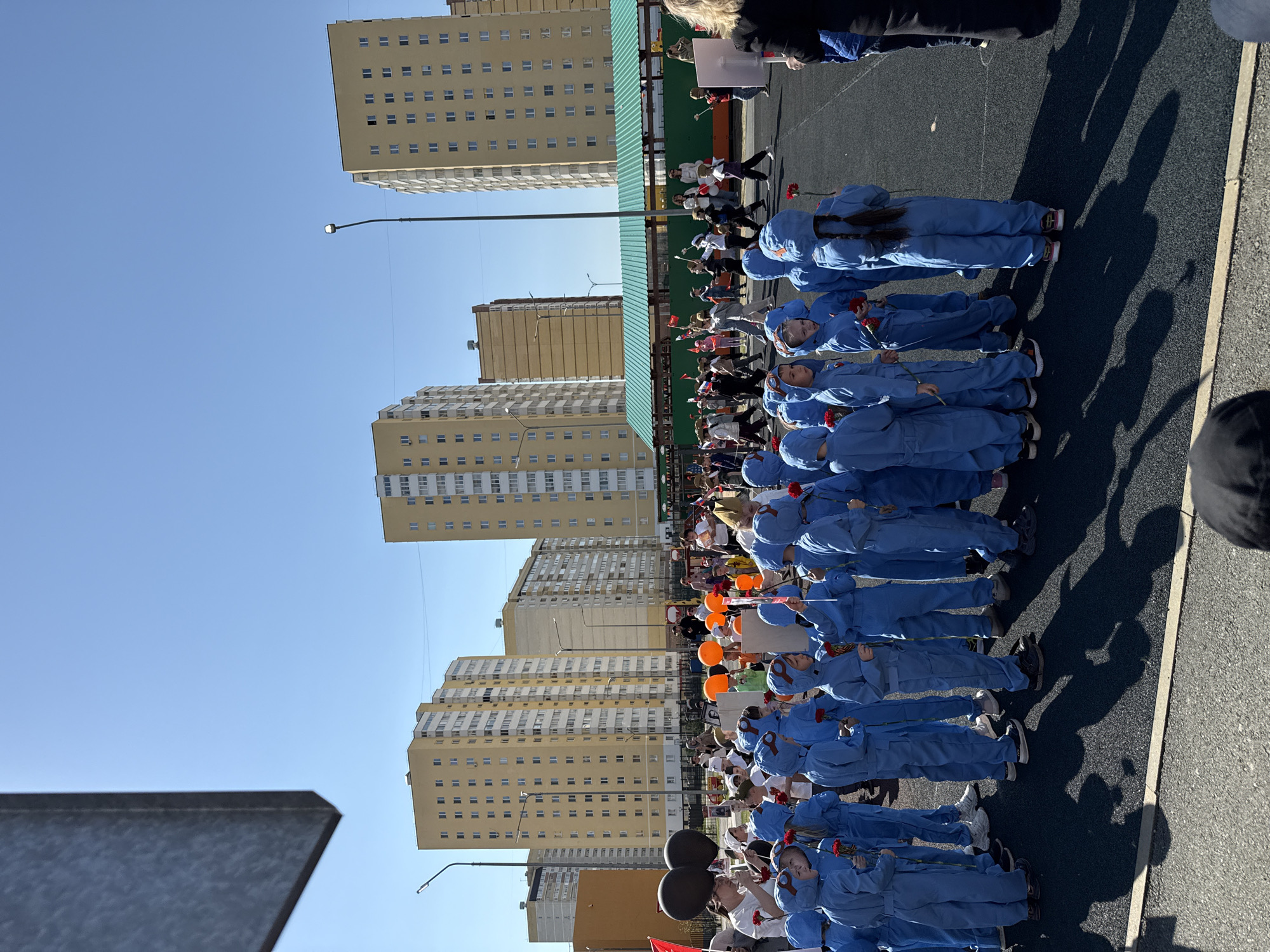 In Tobolsk, Russia, a row of children dressed in blue uniforms lie on a paved surface, each holding a red flower. Tall apartment buildings are visible in the background under a clear blue sky. Behind the children is a crowd of people and orange and black balloons. A street light is also visible. The children are lying flat on their backs, forming a line on what appears to be a street or public square.