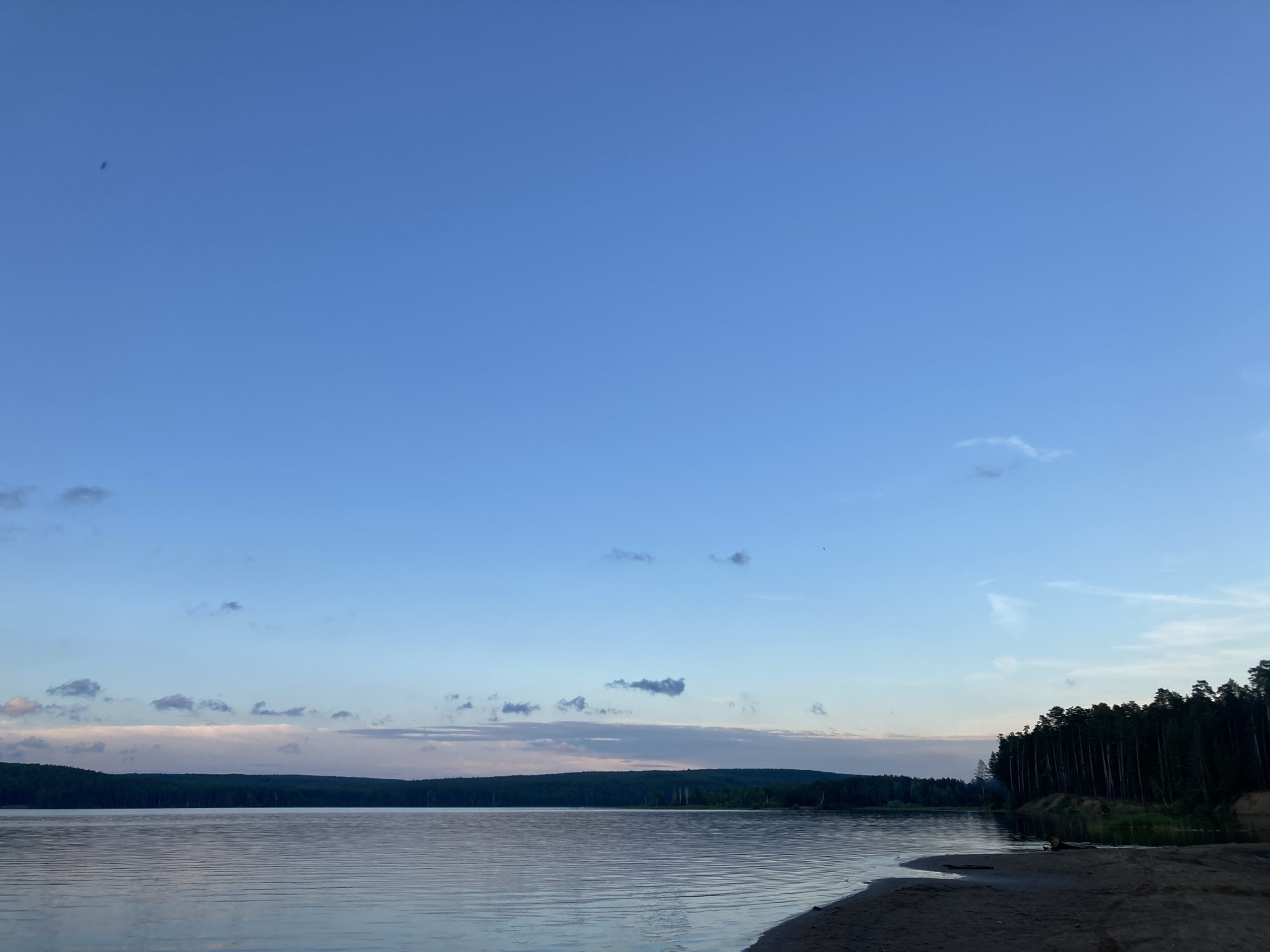 The image depicts a tranquil, wide shot of a body of water, likely a river or lake, under a vast expanse of pale blue sky with scattered, wispy clouds. The water's surface is calm, reflecting the subtle colors of the sky and horizon. Along the far bank, a dense, dark green treeline stretches horizontally, suggesting a natural landscape. On the right side of the foreground, a sandy shore slopes gently towards the water. The overall scene conveys a sense of natural beauty and peacefulness, characteristic of outdoor recreational areas. This landscape is consistent with the natural surroundings found in and around Chaikovsky, Russia, a city known for its picturesque riverbanks and forested areas along the Kama River. There are no people or animals visible in the image. The lighting suggests it is either early morning or late evening, during the twilight hours.