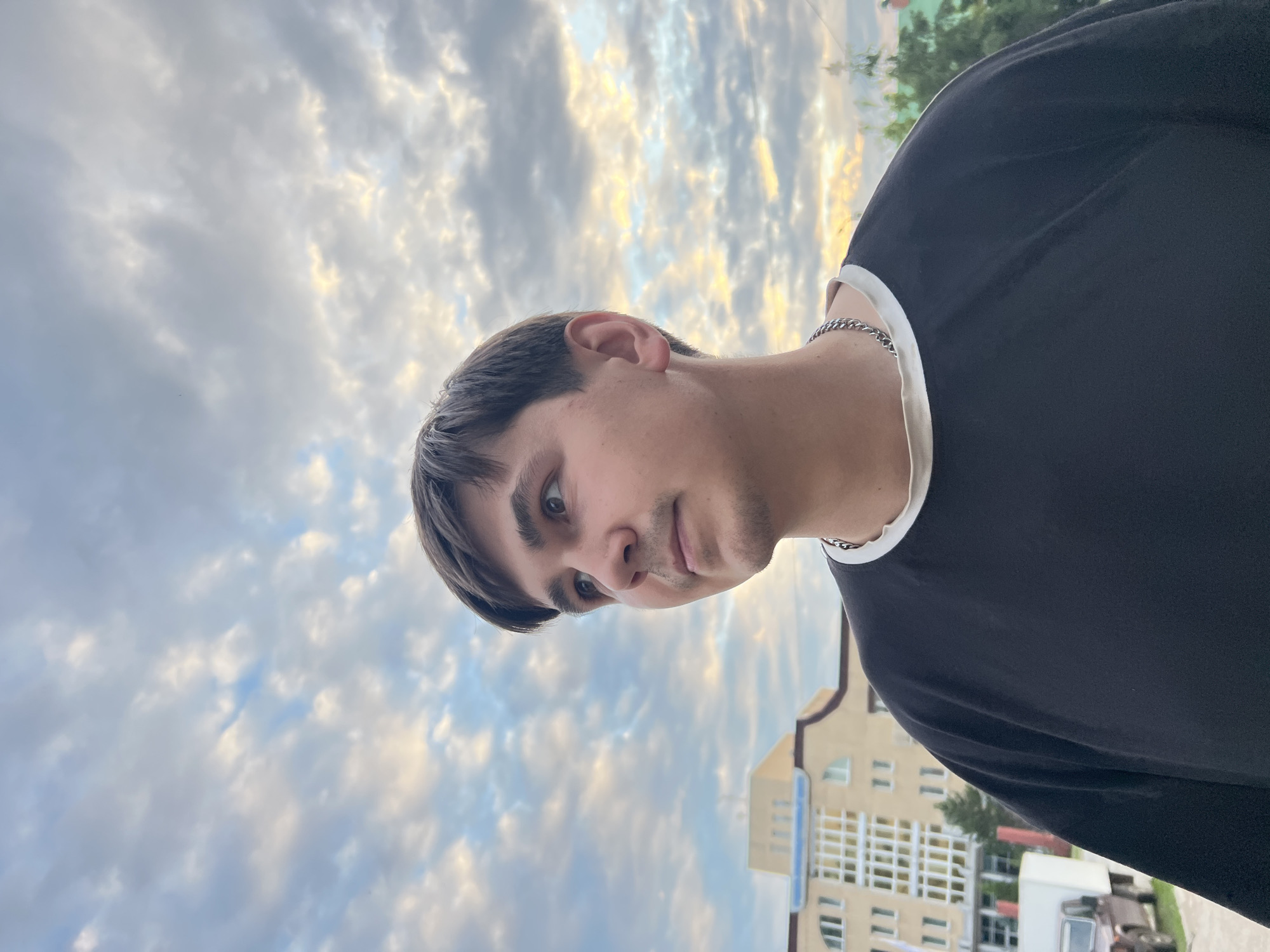 A young man with brown hair and brown eyes is captured in a close-up shot outdoors. He is wearing a black t-shirt with a white trim around the collar and a silver chain necklace. The man is looking to his left, with a thoughtful expression. In the background, a beige building with numerous windows is visible, alongside some green trees and a vehicle. The sky above is filled with dynamic clouds, suggesting it might be late afternoon or early evening. The scene appears to be set in a residential or urban area.