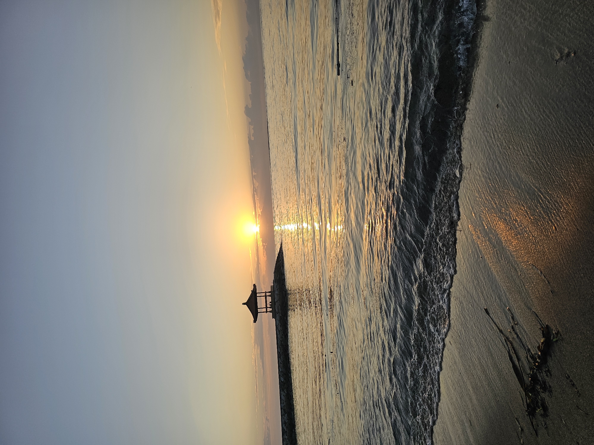 This is a vertical, eye-level, medium shot of a serene sunrise over the ocean at Sanur Beach, Denpasar, Indonesia. The sun is a bright, golden orb, partially obscured by a thin layer of clouds on the horizon. Its reflection shimmers on the calm, rippling surface of the sea, creating a path of light that stretches towards the shore. In the middle distance, a stone pier extends from the left side of the frame into the water, culminating in a traditional Balinese pavilion, silhouetted against the bright sky. Two figures are visible within the pavilion. The foreground is dominated by the wet sand of the beach, with gentle waves lapping at the shore, leaving a foamy edge. Some dark seaweed is scattered on the sand near the water. The sky transitions from a pale blue at the top to a warm yellow and orange near the horizon, indicating the early morning light. The overall mood of the image is peaceful and tranquil.