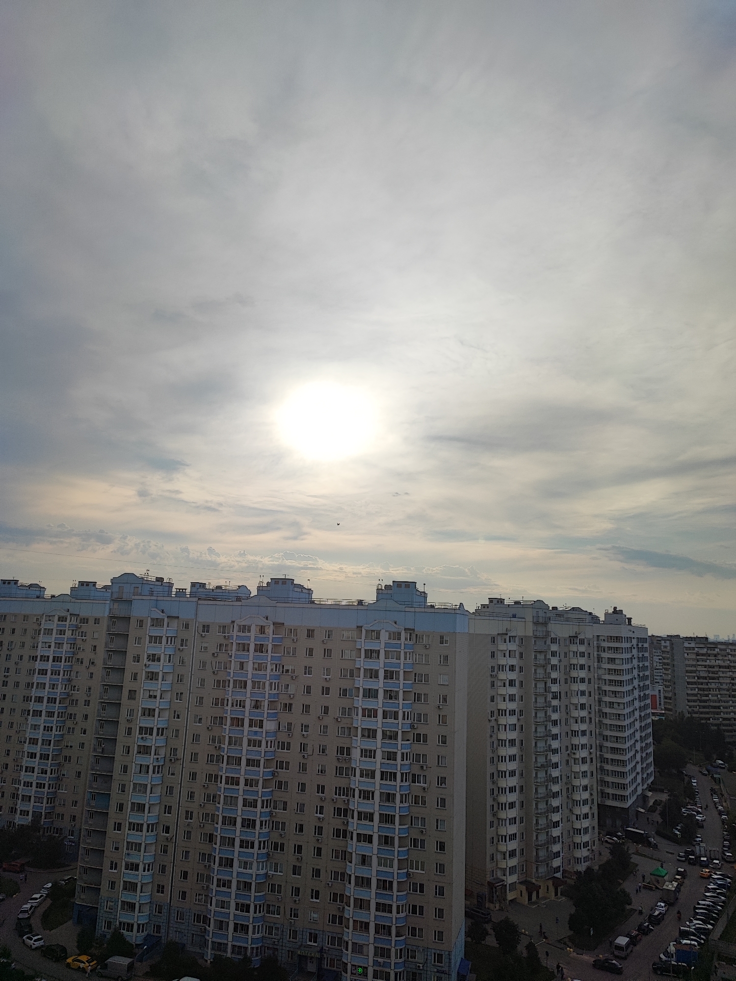 This is a vertical, eye-level, medium-distance shot of a city skyline in Vidnoye, Russia. The foreground is dominated by a row of tall, light-colored apartment buildings with blue accents. The buildings are multi-story with numerous windows and balconies. In the mid-ground, the sun is visible as a bright, hazy orb in a cloud-filled sky. The clouds are a mix of white and gray, with streaks of lighter and darker tones. In the background, more apartment buildings are visible, as well as a street with parked cars. The overall impression is one of a densely populated urban environment under a cloudy sky.
