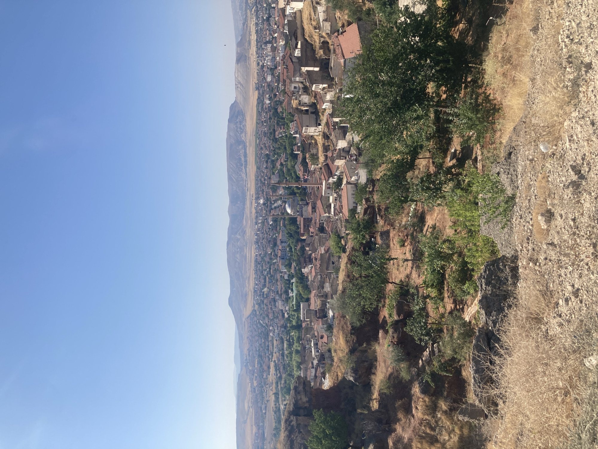 The image shows a panoramic view of Avanos İlçe Merkezi, Turkey, a town characterized by its dense urban fabric nestled within a valley. The foreground is dominated by a dry, rocky landscape with sparse vegetation, featuring reddish-brown soil and scattered rocks. A few small trees and shrubs are visible, their foliage appearing somewhat dry and yellowed, consistent with a warm, arid climate.
In the mid-ground, the town of Avanos unfolds. Numerous buildings with terracotta-colored roofs are tightly packed together, forming a mosaic of urban development. Many structures appear to be traditional, with stone or plaster walls. Distinctive elements of the town's architecture, such as minarets, are visible, suggesting the presence of mosques. The overall impression is one of an old, established settlement integrated into the natural terrain.
Beyond the town, a vast expanse of rolling hills and distant mountains stretches across the horizon. The landscape is arid and predominantly brown and yellow, with some sparse vegetation. The sky is a clear, bright blue, indicating a sunny day. The lighting suggests the photograph was taken during daylight hours, likely in the morning or afternoon. No people are visible in the image. The scene is static, offering a still representation of the town and its surrounding environment.