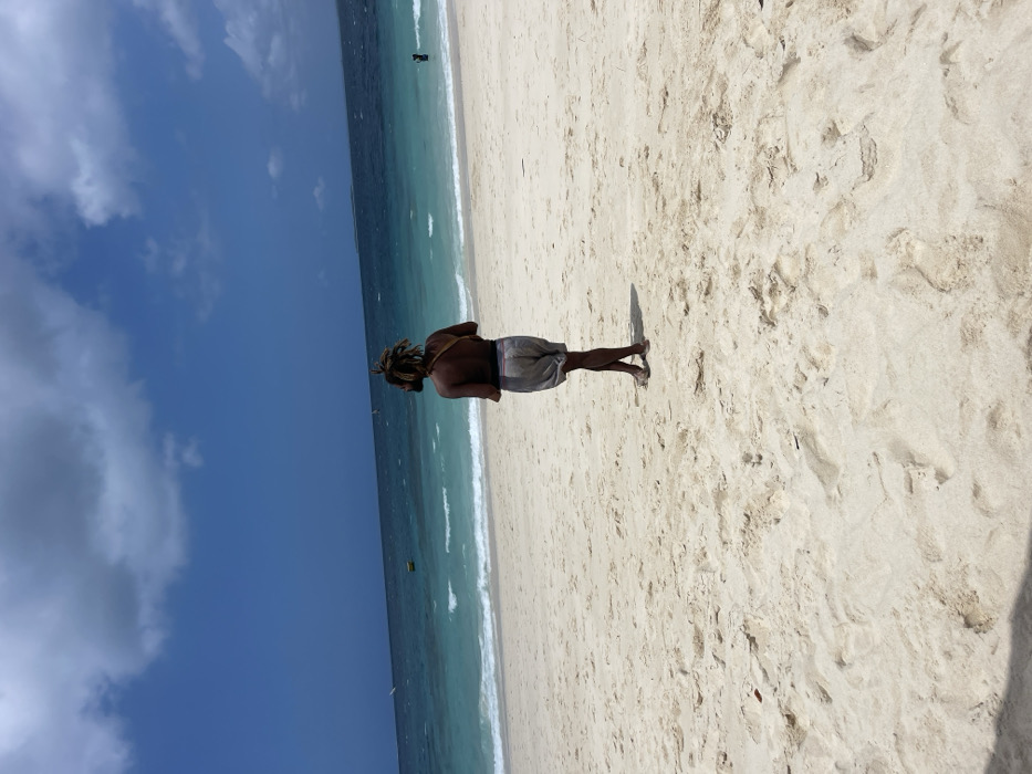 The image depicts a beach scene in Zanzibar, Tanzania. A lone individual is positioned centrally on light-colored sand, viewed from behind, facing the ocean. This adult figure has dark skin, brown dreadlocks, wears grey shorts, and no shirt, with a dark strap visible over their left shoulder. Their legs are crossed at the ankles. The ocean displays varying shades of blue and turquoise, with moderate waves breaking near the shore. Within the water, a yellow buoy is visible in the mid-distance to the left, two white-sailed vessels are further out on the left, and a person is partially submerged on the far right. The sky above is blue with scattered white and grey clouds.