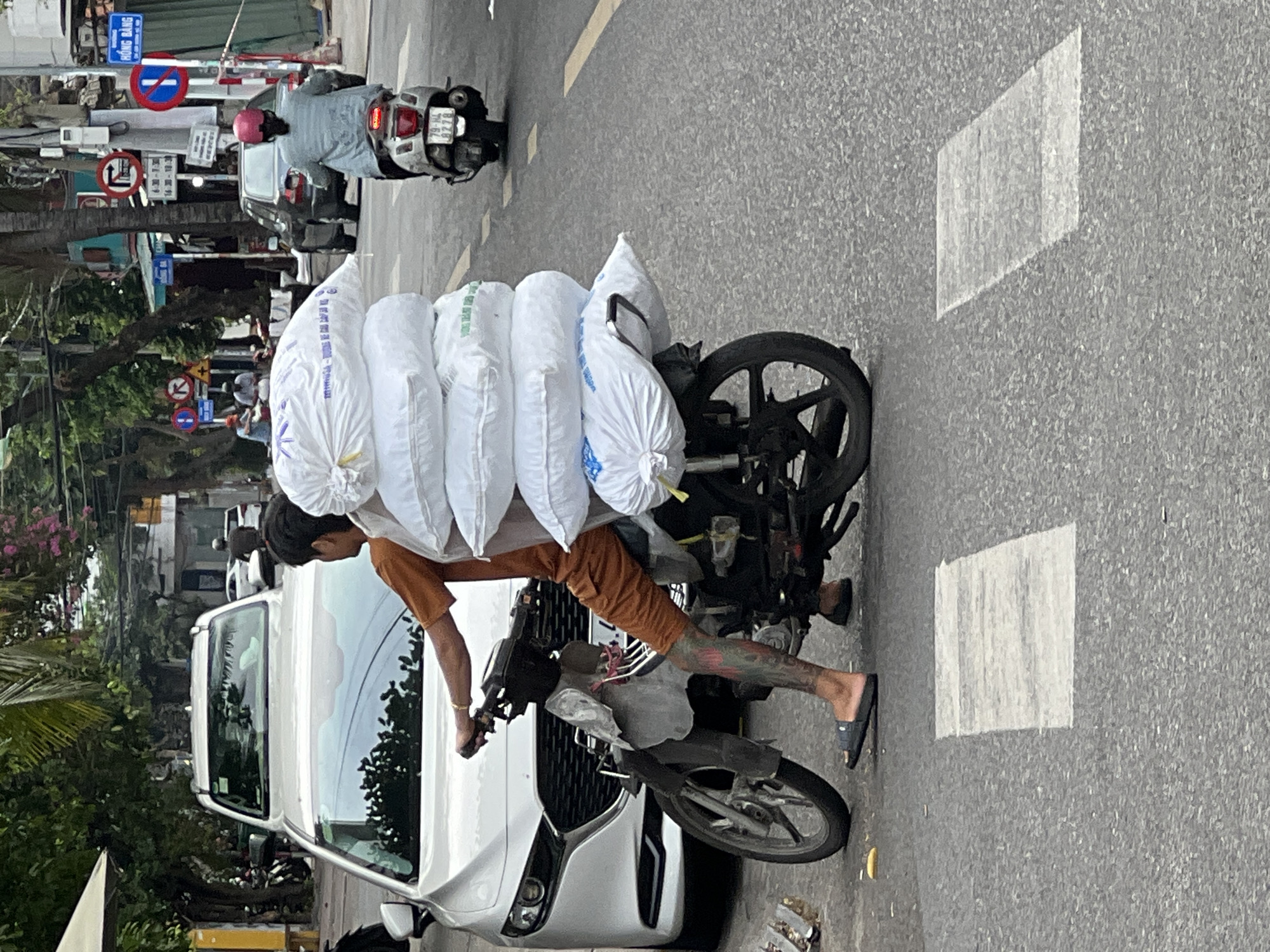 A man is riding a motorcycle on a street in Nha Trang, Vietnam, carrying a large stack of white bags on the back of his vehicle. The man is wearing an orange t-shirt and orange shorts, and has tattoos on his legs. He is wearing dark-colored flip-flops. The motorcycle is a dark color and appears to be a scooter. The bags are large, white, and appear to be filled with a soft material. The motorcycle is in motion, and the man is looking forward. In the background, there is a white car and other vehicles, as well as buildings and trees. The street has white crosswalk markings. The image appears to be taken from a slightly elevated position, looking down at the scene.