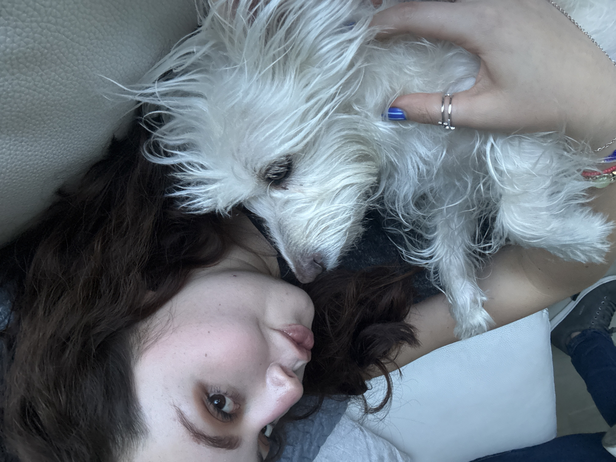 A woman with dark curly hair and light skin lies on a white cushion, possibly a sofa, with a small white dog resting on her chest. The woman's face is partially visible, showing her left eye, nose, and lips. Her right hand, adorned with a ring and blue nail polish, is gently placed on the dog. The dog has long, shaggy white fur. Part of the woman's leg is visible in the lower right corner, wearing dark blue jeans and gray shoes. The location in the city of Santa Clara Ocoyucan, Mexico cannot be determined from the image.