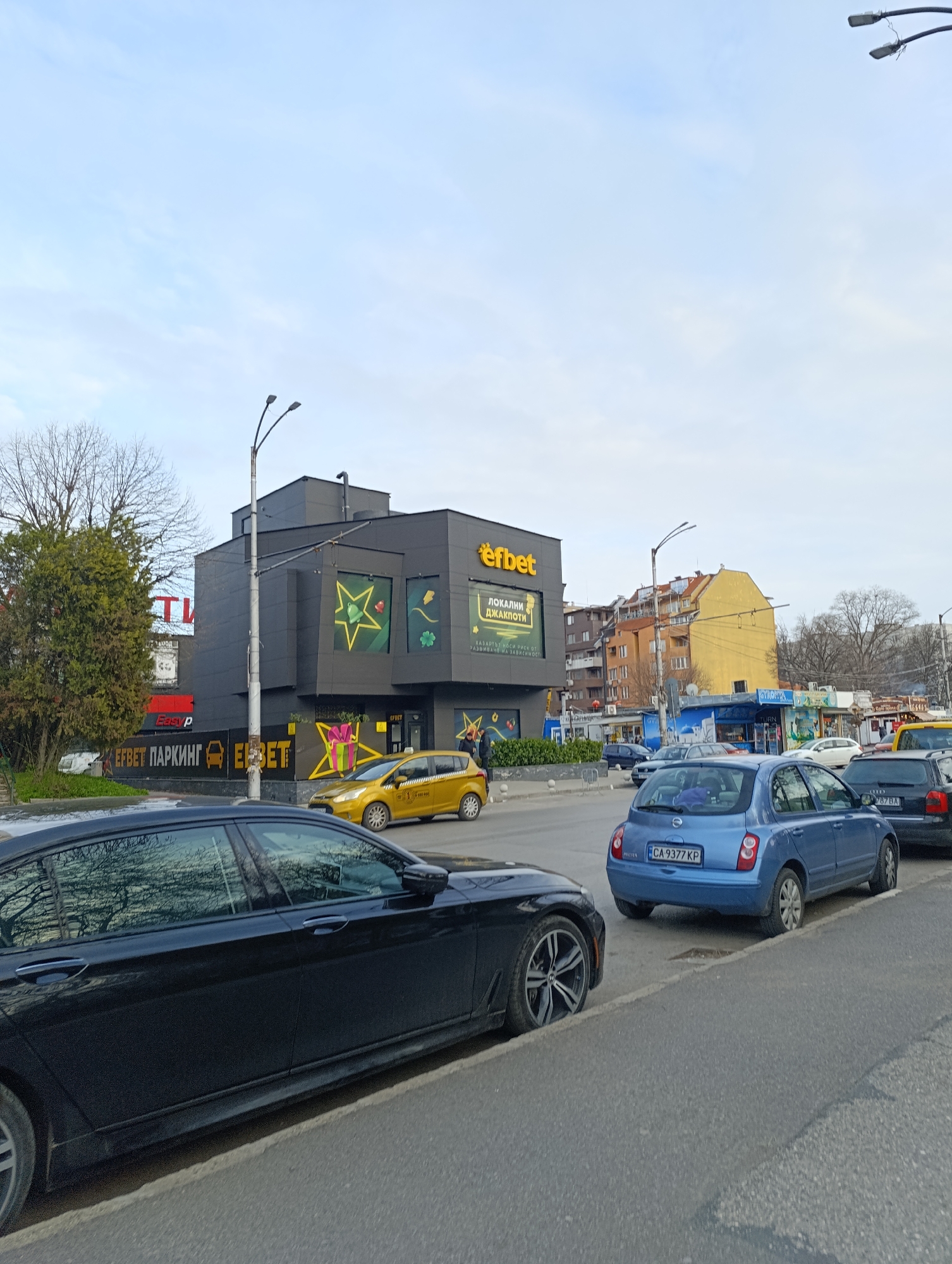 Here's a concise, forensic description of the image:
**Location:** Sofia, Bulgaria, a street scene showing an EFbet gambling establishment.
**Subjects and Objects:**
* A modern, black, multi-story EFbet building with large star and cloverleaf decals. There is text on the building, "efbet" and "ЛОКАЛНИ ДЖАКПОТИ".
* Multiple cars are parked along the curb: a black sedan in the foreground, a blue Nissan Micra, a black sedan behind it, and a yellow taxi near the EFbet building.
* There are pedestrians in the background near the EFbet building.
* Trees, utility poles, and other buildings are visible in the background, giving context to the urban location.
* There are smaller retail kiosks situated near the bus stop behind the parked cars.
**Environment:**
* The sky is overcast.
* The scene is set on a city street with standard traffic and parking.
* The architecture includes a mix of modern and older buildings.
**Action/Interaction:**
* Traffic is present on the street.
* People are walking near the gambling establishment.
* Cars are parked along the street.