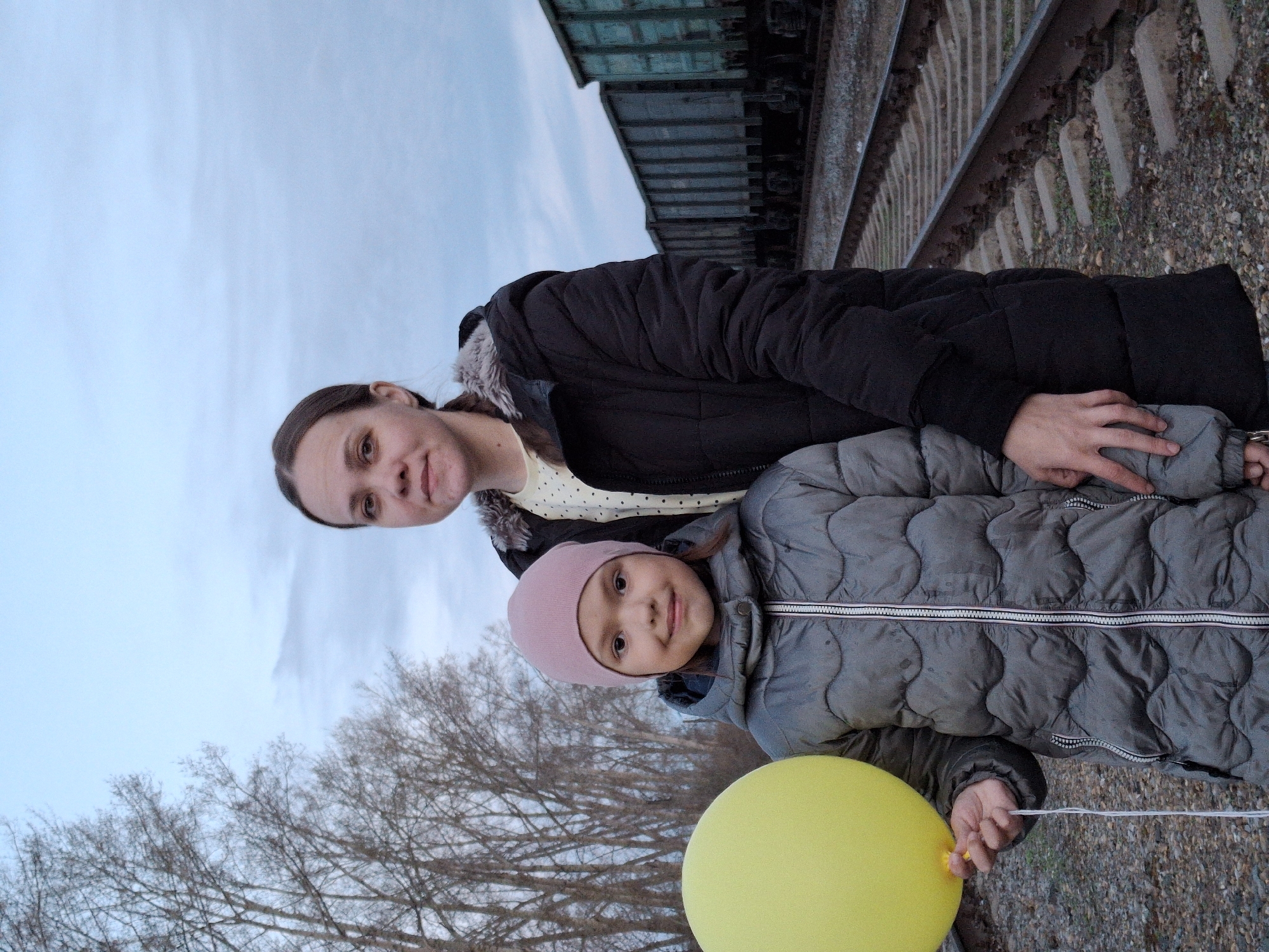 This outdoor portrait features an adult woman and a young girl standing beside a railway line. The scene is set in what appears to be late autumn or early spring in Chaikovsky, Russia, characterized by bare trees and an overcast sky.
The adult woman is positioned slightly behind and to the left of the child. She has long, dark hair and is dressed in a dark winter jacket with a light-colored, polka-dotted top visible underneath. Her left hand is resting on the child's shoulder. She looks directly at the camera with a calm expression.
The young girl stands prominently in the foreground, wearing a light pink knitted beanie and a grey puffer jacket. She holds a bright yellow balloon by its string in her right hand. The girl is smiling gently towards the camera.
To the right of the subjects, railway tracks are clearly visible, with concrete sleepers embedded in gravel ballast. A dark green train car, possibly a freight or container car, is partially visible parked on the tracks in the background. The sky is a uniform light blue-grey, suggesting an overcast day. To the left, a dense network of bare tree branches frames the scene against the sky, indicating a time of year when leaves have fallen. The overall mood is quiet and possibly cool, given the subjects' outerwear.
