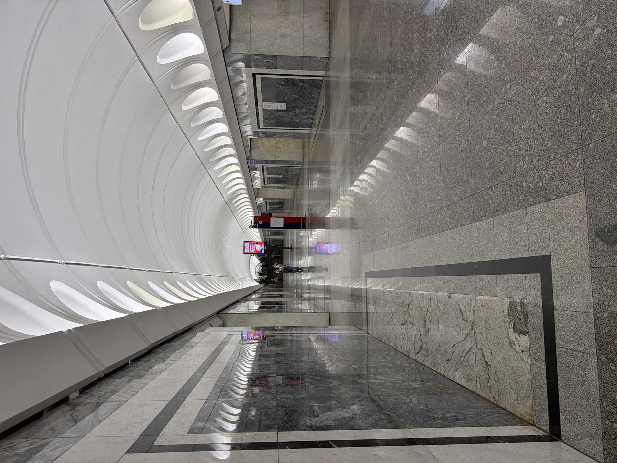 This image captures the interior of a Moscow Metro station. The station features a grand, arched ceiling with integrated lighting, creating a modern and spacious atmosphere. The walls are clad in polished marble with a geometric pattern, reflecting the light from the ceiling and contributing to the station's elegant aesthetic. The floor is also made of polished stone tiles, with a reflective surface that mirrors the architectural elements. Digital screens are visible on the walls, displaying advertisements or information. In the distance, a few individuals are seen walking along the platform, one person in the mid-ground is walking away from the camera. The overall scene suggests a well-maintained and visually impressive public space typical of the Moscow Metro system.