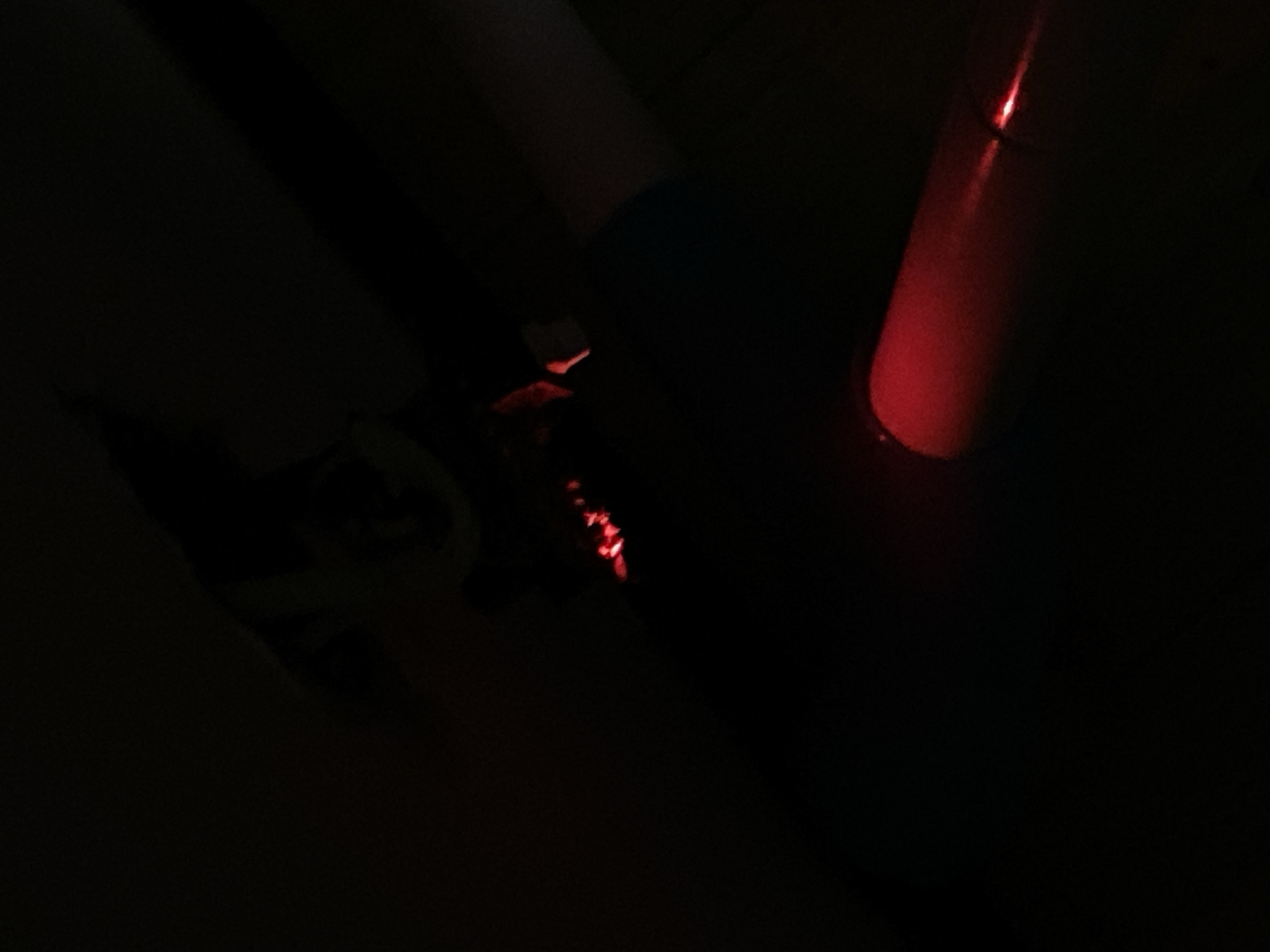 The scene is set in Chaikovsky, Russia, under extremely low-light conditions, rendering most of the environment indiscernible. The primary visible subjects are two distinct sources of light. In the upper-middle section of the frame, an irregular cluster of approximately six to eight intensely glowing orange-red points is observed, consistent with active embers or a smoldering object. Below and to the right of the frame's center, a cylindrical object emits a concentrated, continuous red light from its end and appears to reflect this light along a portion of its surface, indicating a smooth or reflective material. The surrounding areas are enveloped in profound darkness, precluding the identification of any specific background objects, people, actions, or interactions.