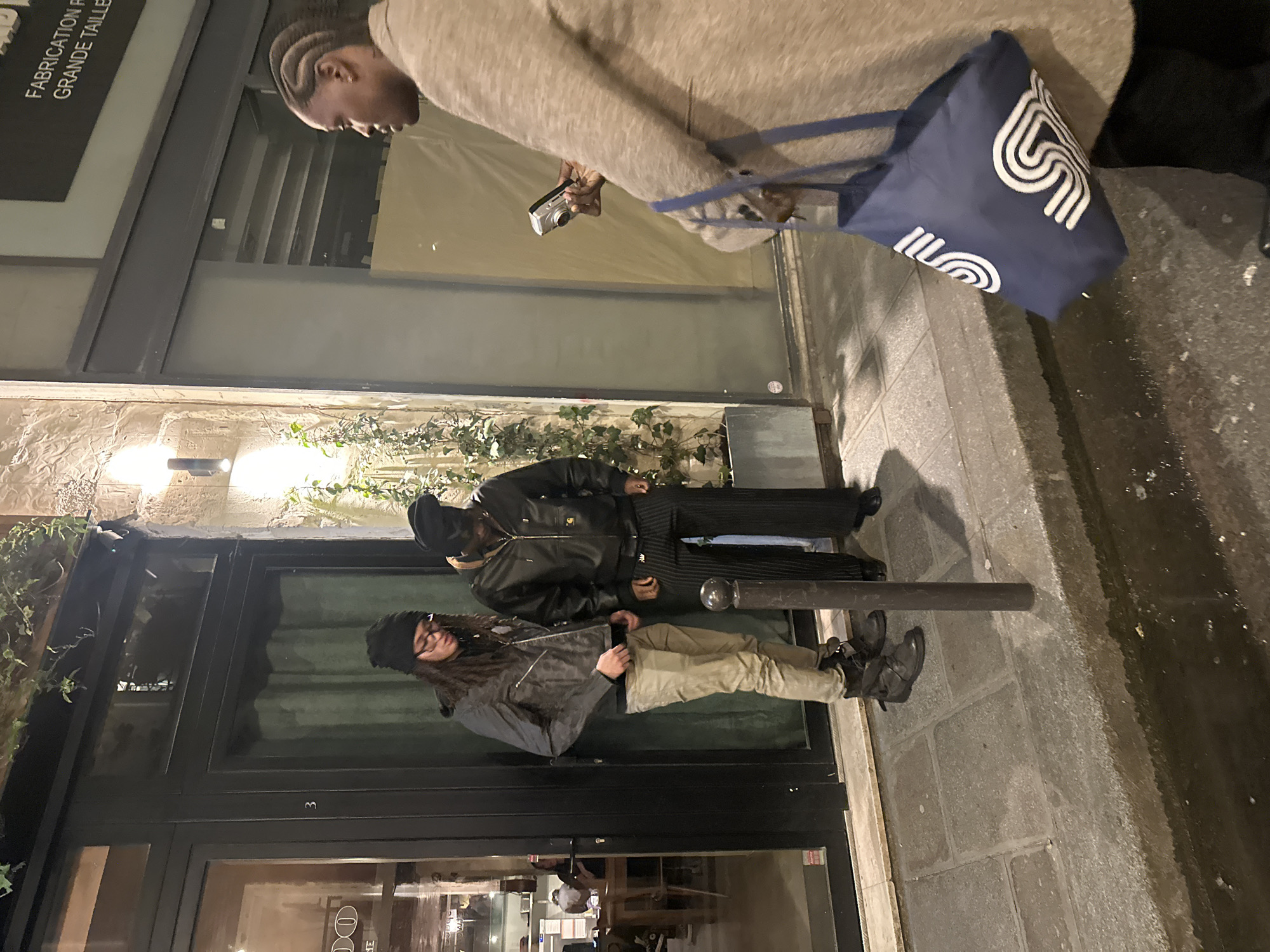 The scene depicts a nighttime street in Paris, France, featuring three individuals on a paved sidewalk next to a building facade. The building has a rough-textured stone wall on the left and a smoother, lighter section on the right, punctuated by dark-framed glass doors and windows. One door bears the number "3". Above a large window to the right, a sign with white text on a dark background reads "FABRICATION GRANDE TAILLE". Ivy grows in planters against the stone wall, illuminated by two modern, vertical wall-mounted light fixtures.
On the left, a person with long dark hair, wearing a black beanie, eyeglasses, a dark grey patterned jacket, tan pants, and dark, multi-strapped boots, stands facing downwards. Next to them, a second person wears a black beanie, a black leather bomber jacket with a white "Carhartt" logo on the left chest, dark pinstriped wide-leg pants, and dark shoes; this individual also looks downwards. Both are positioned close to the building's entrance.
To the right, a third person, primarily visible from the mid-torso to the knees, wears a light brown, long-sleeved coat and dark pants. This individual has dark braided hair, holds a silver compact digital camera in their right hand, pointed towards the two individuals on the left, and carries a dark blue tote bag with white stylized "S" or wave logos on their left arm. A dark metal bollard stands on the sidewalk between the two groups. The sidewalk is paved with rectangular stone slabs, and a narrow, dark puddle is visible along the curb in the immediate foreground.