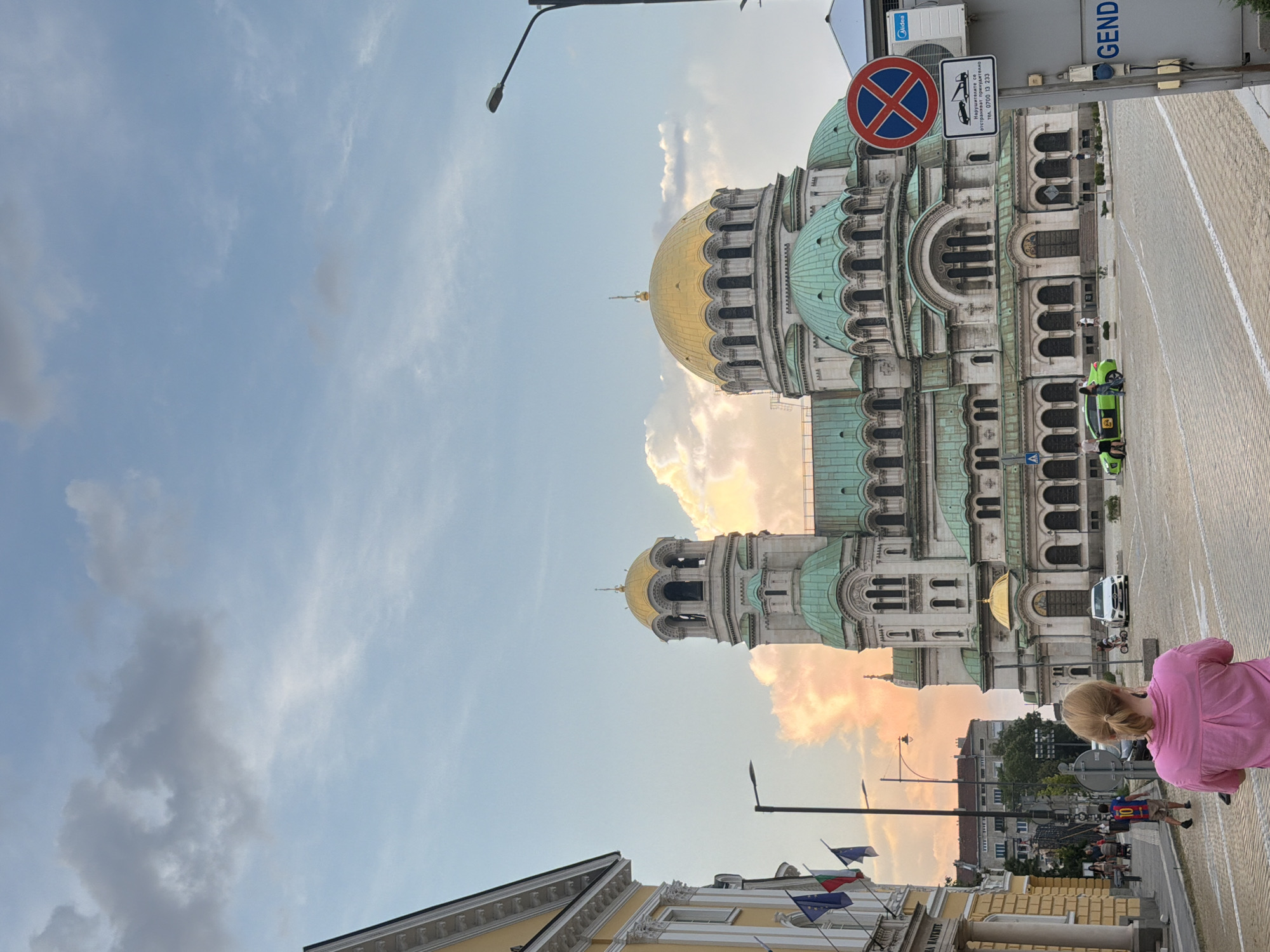 The image captures a wide view of Alexander Nevsky Cathedral in Sofia, Bulgaria, at dusk. The cathedral, a prominent landmark with golden domes and green roofs, dominates the center of the frame. In the foreground, a woman with blonde hair pulled back in a ponytail, wearing a pink shirt, faces away from the camera, looking towards the cathedral. The street in front of the cathedral is paved with cobblestones. Several people are visible in the middle ground, including a man wearing a soccer jersey with the number 10, walking away from the camera, and a green taxi with passengers. Cars and a child on a bicycle are also present in the plaza. To the left of the cathedral, a yellow building with flags is visible. A street light and a "no parking" sign are positioned to the right of the cathedral. The sky is a mix of blue and cloudy, with hints of sunset colors.