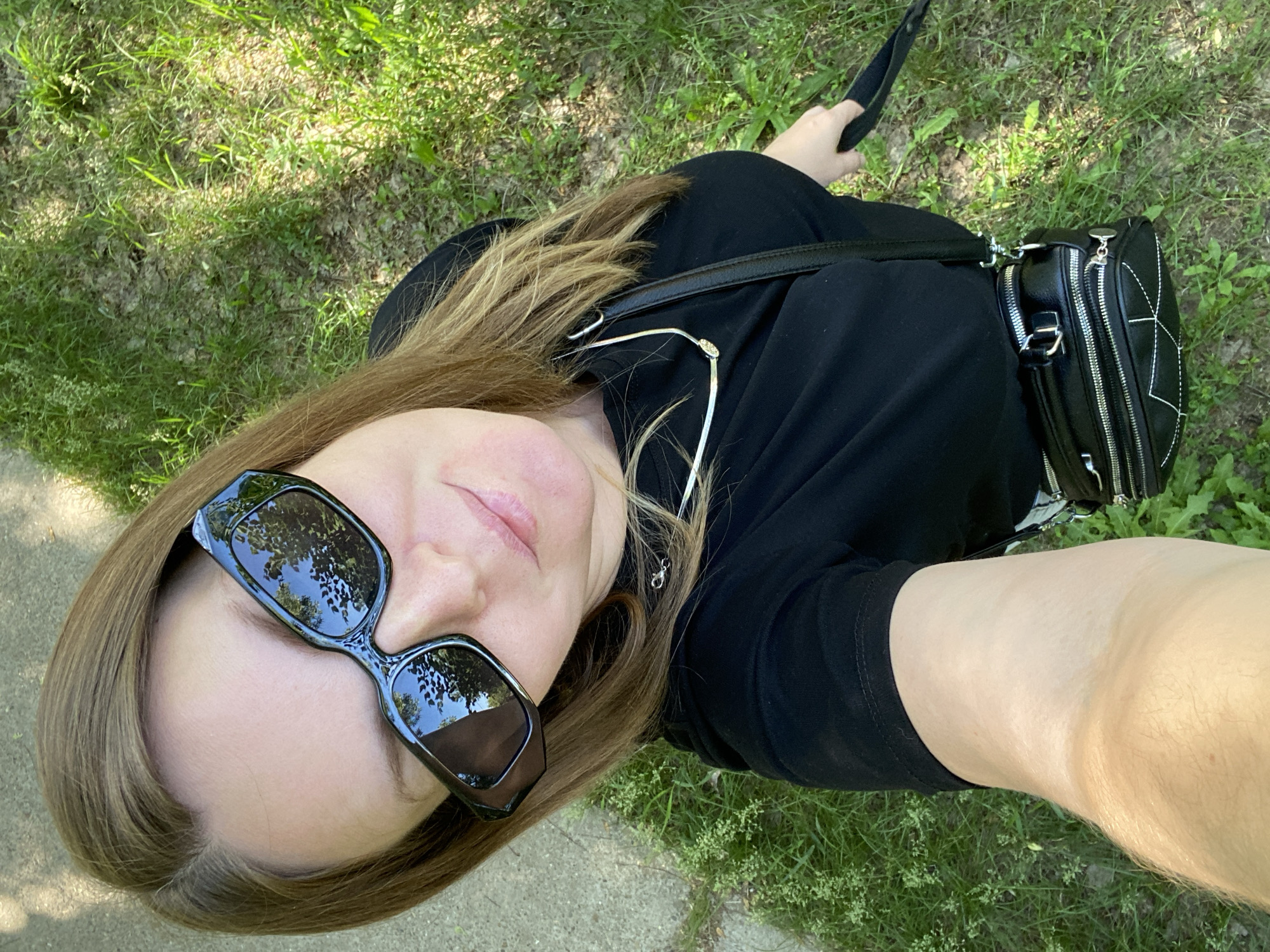 A woman is taking a selfie outdoors on a sunny day. She is wearing black clothing, sunglasses, and a necklace. She has a black purse with zippers and stitching across her body. Her arm is extended, holding the camera. The background shows grass and some pavement or sidewalk. The location is not specified in the image, but it is requested to be Noviy, Russia.