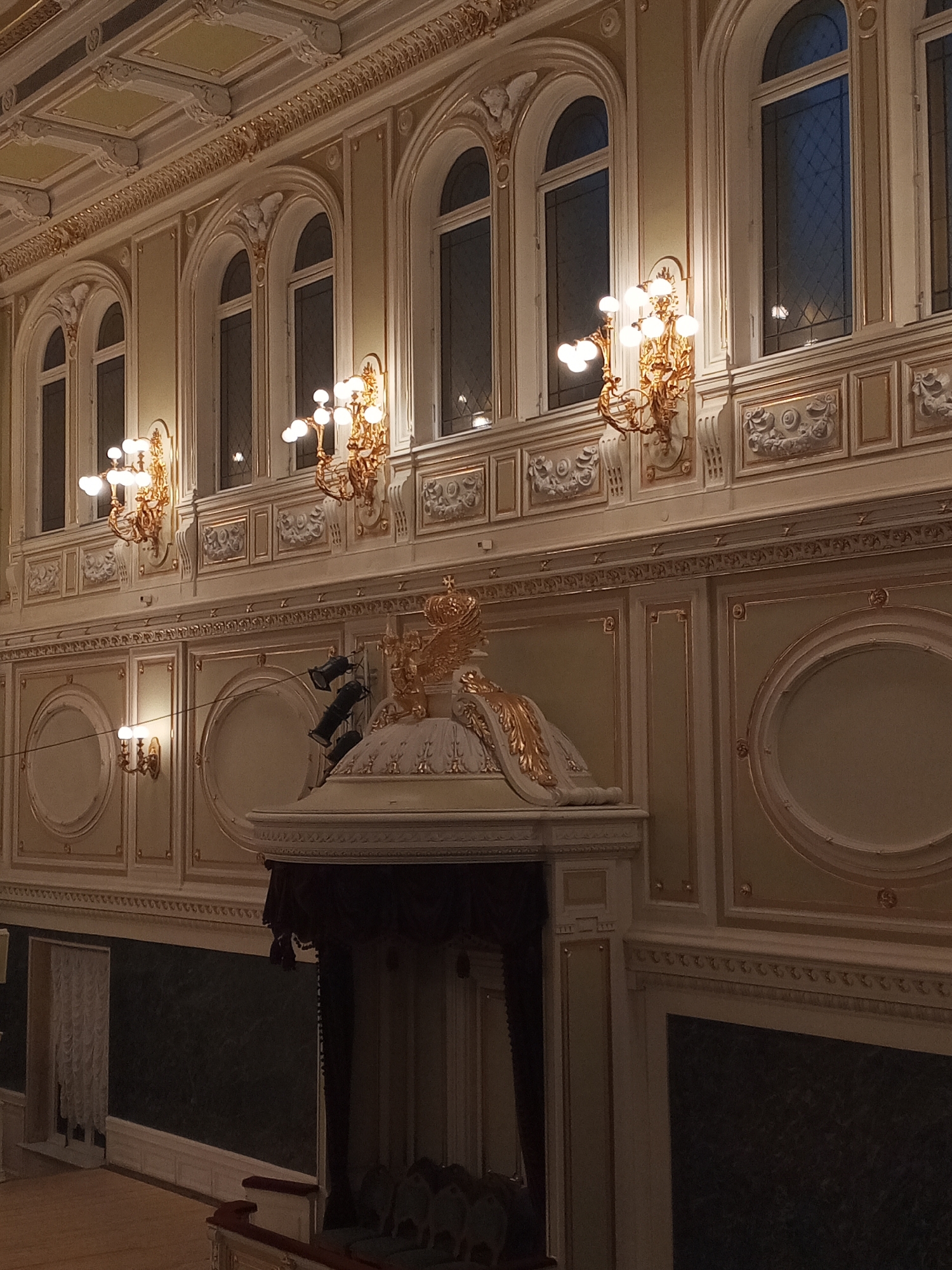 The image shows the interior of a theater. The architecture is ornate, featuring cream-colored walls with gold detailing. There are several tall arched windows along the upper portion of the wall. Wall sconces with multiple lights are placed between the windows. Below the windows, the wall has decorative panels and circular design elements. A prominent decorative structure with a gold eagle and draped fabric is in the foreground. The lower portion of the wall is dark green. Seating is visible in the background. The location is likely in Saint Petersburg, Russia, given the style of architecture.