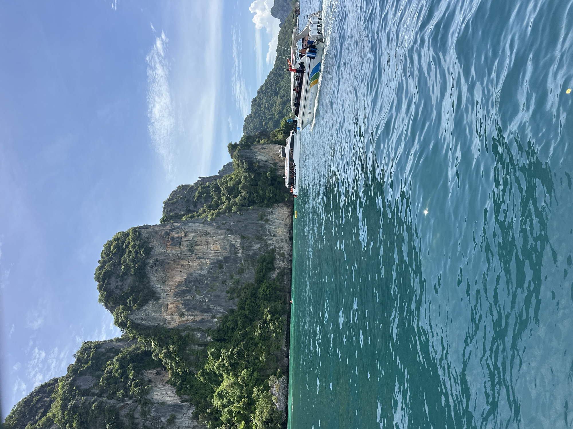 In Ao Nang, Thailand, a speedboat with several passengers speeds through turquoise waters. Towering limestone cliffs covered in lush green vegetation rise dramatically from the sea in the background, under a bright blue sky dotted with white clouds. Another larger boat, labeled "SAWANU TRAVEL," is visible in the distance. The water is choppy, with whitecaps and reflections of the sky and cliffs. A person in a red life vest appears to be standing on the back of the moving speedboat, interacting with other passengers.