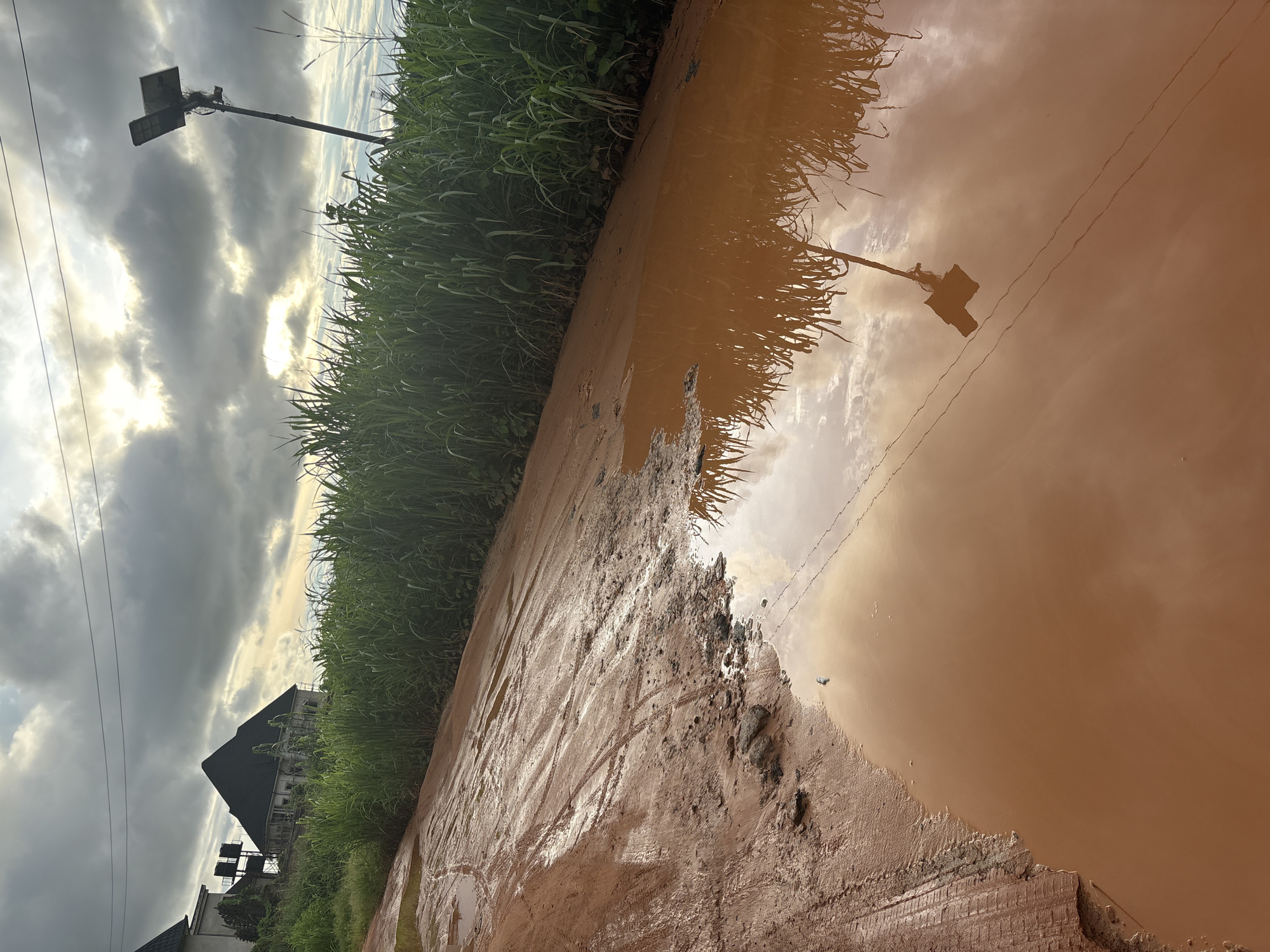 An overcast sky with patches of sunlight looms over a muddy road in Obosi, Nigeria. The road is partially submerged in a large puddle, reflecting the sky, tall grass, a solar-powered street light, and overhead power lines. Tire tracks are visible in the mud. In the background, a partially constructed building with a dark, pyramid-shaped roof and water tanks on its side stands amidst dense green vegetation.