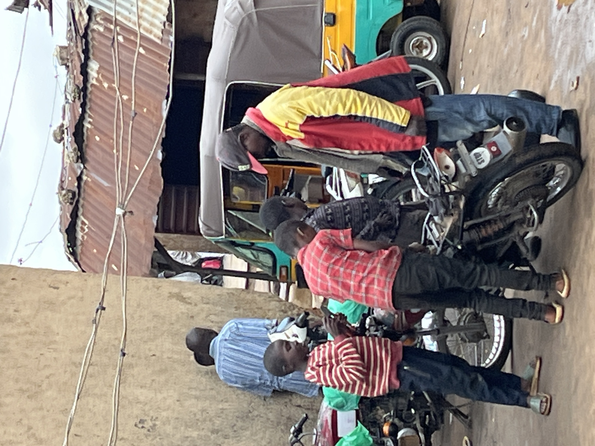 In Gura Top, Nigeria, a group of individuals are gathered in a street scene. In the foreground, two young boys stand next to a motorcycle. The boy on the left wears a red and white striped long-sleeved shirt and dark blue pants, with sandals on his feet. He is holding a dark object, possibly a phone. The boy on the right wears a red and black plaid short-sleeved shirt and dark pants, also in sandals. Behind them, an adult male in a red and yellow jacket and a dark baseball cap looks downwards. Further back, another adult, partially obscured, wears a blue and white striped shirt. The environment is characterized by a building with a rough, beige wall on the left, and a corrugated metal roof on a structure to the right. A three-wheeled vehicle, possibly an auto-rickshaw, is visible behind the people. Motorcycles are parked around the area, suggesting a busy marketplace or transportation hub. The ground is a rough, reddish-brown surface.