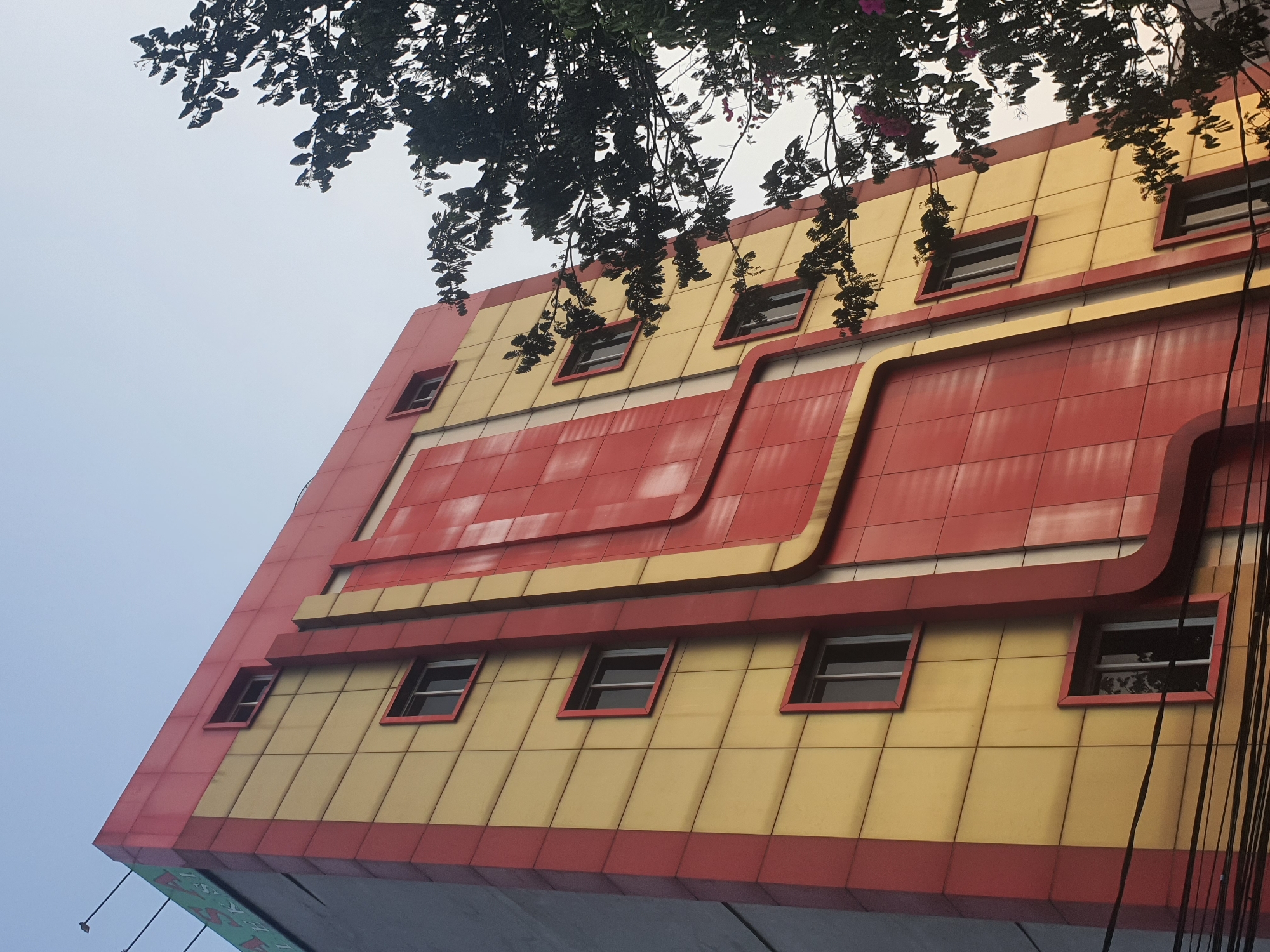 This is a low-angle exterior shot of a multi-story building in West Jakarta, Indonesia. The building features a striking design with alternating bands of bright yellow and red panels. Red trim outlines the yellow panels and frames several windows. A prominent red, curved vertical feature runs down the center of the facade. Trees with dark green foliage and some pink flowers partially obscure the right side of the building. The sky above is a pale, overcast blue. Several dark electrical wires are visible in the foreground, running horizontally across the lower portion of the image. The building appears to be a commercial establishment, possibly a hotel or office building, given its size and prominent signage that is partially visible at the top left of the image, reading "ASA" and "LEKSI".